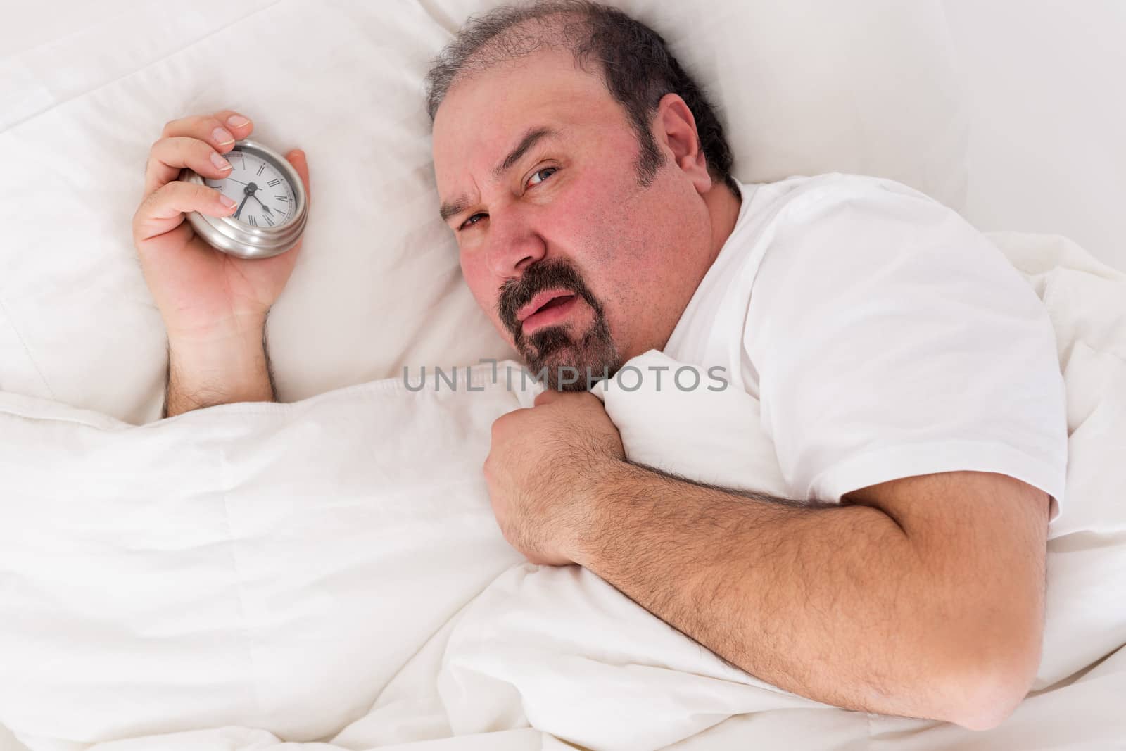 Irascible man glaring balefully at the camera as he lies in bed holding the alarm clock in his hand after a sleepless night wanting to remain in bed until noon