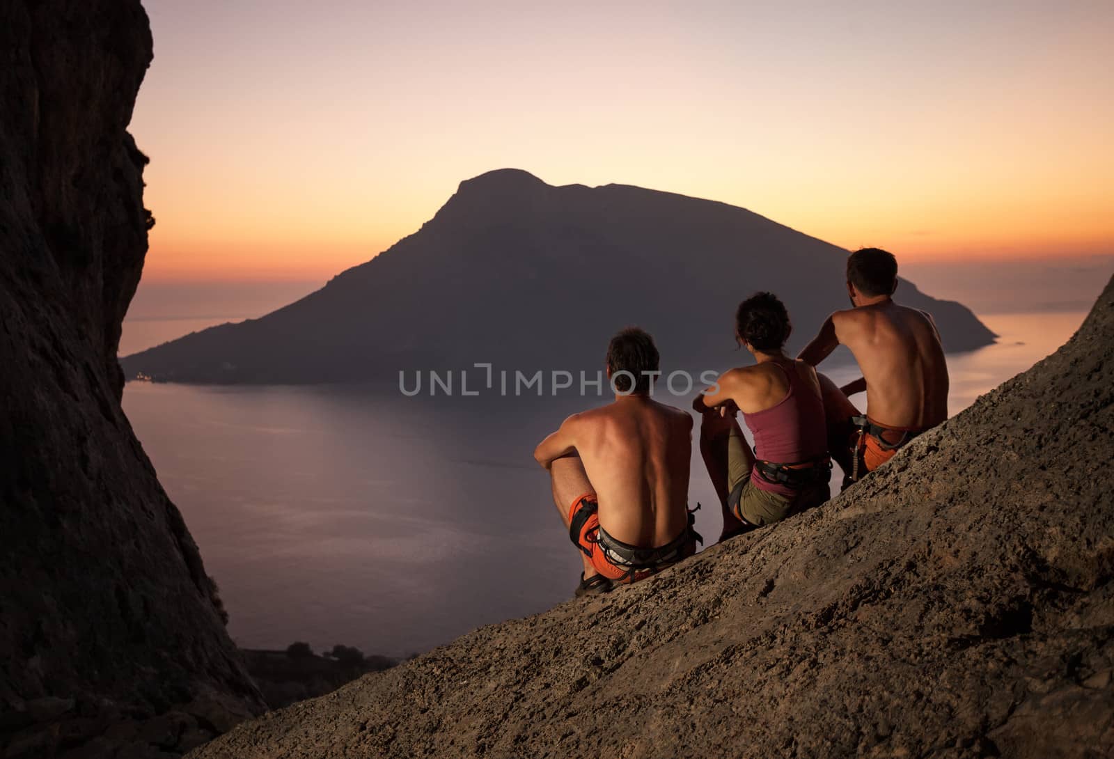 Three rock climbers wearing safety harness having rest at sunset. With picturesque view of Telendos Island in front. Kalymnos Island, Greece.