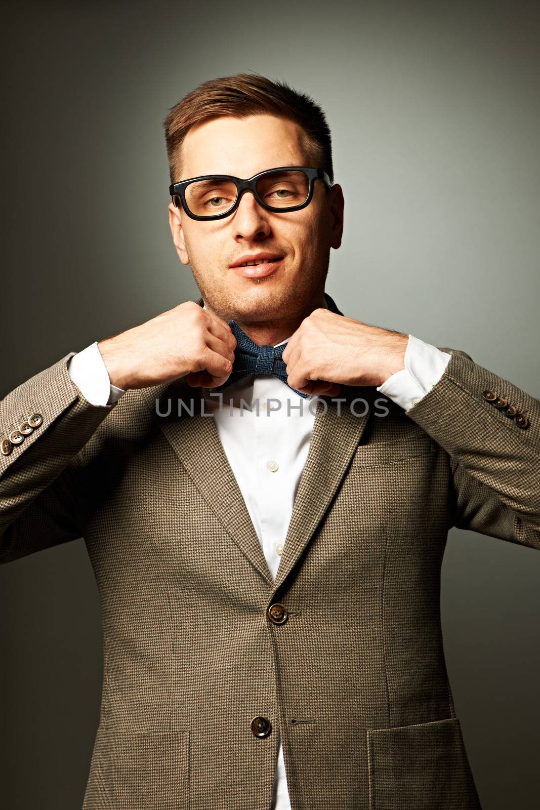 Confident nerd in eyeglasses adjusting his bow-tie against grey background
