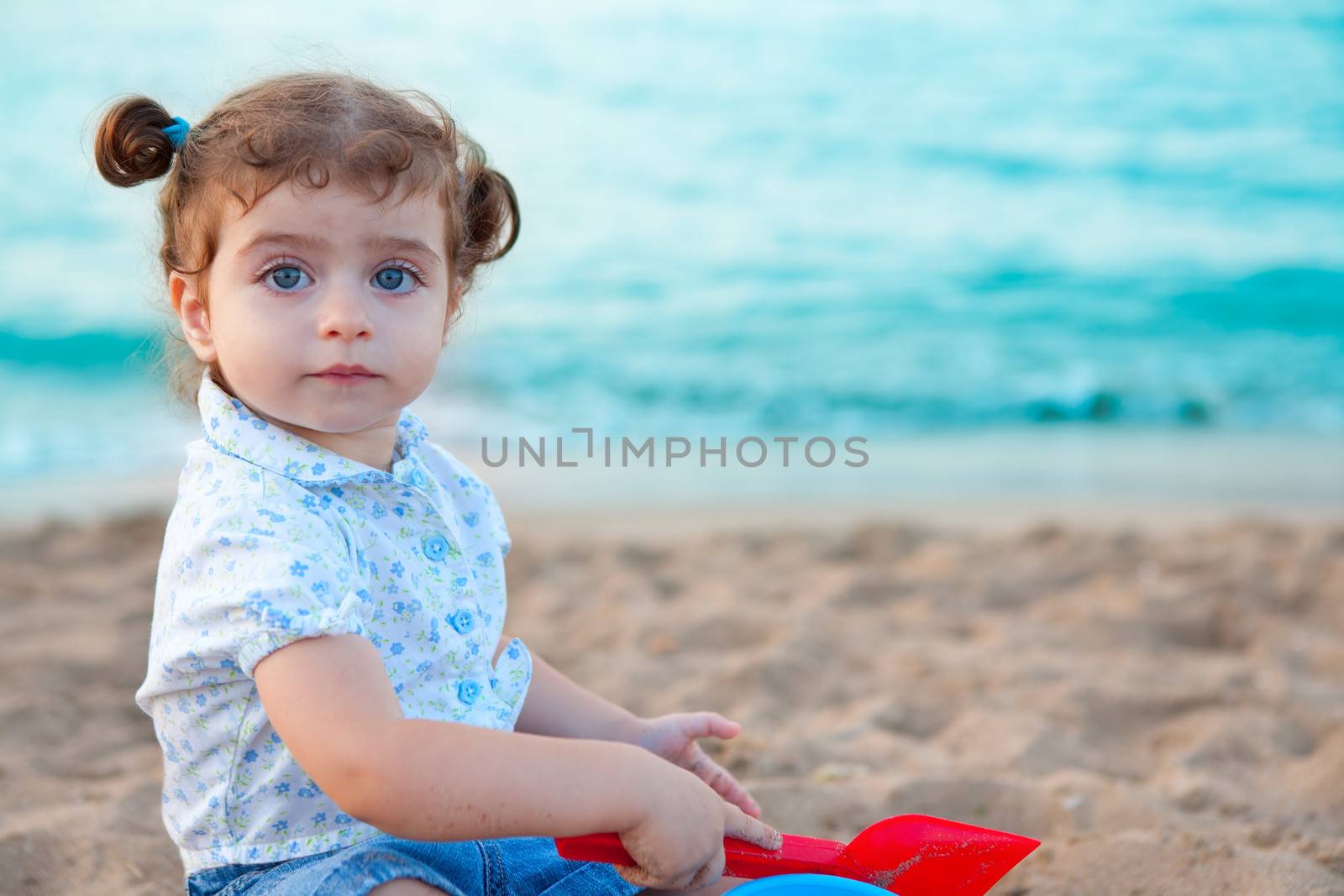 Blu eyes brunette toddler girl playing with sand in beach by lunamarina