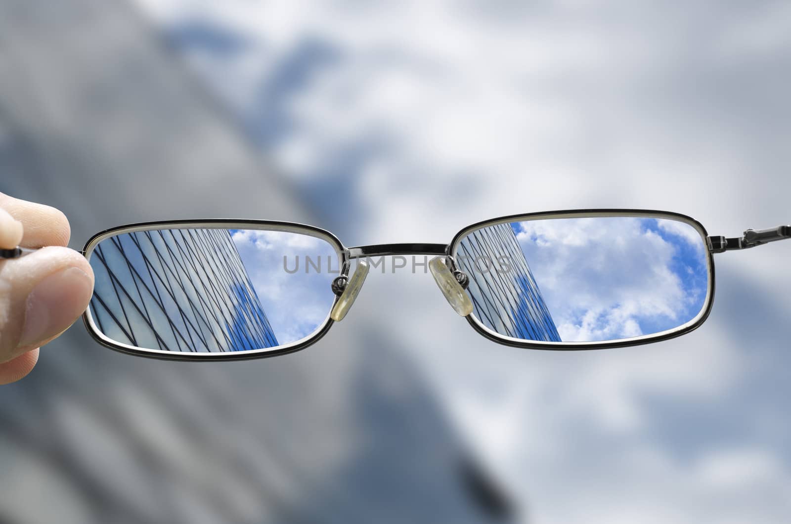 out of focus glass business building with sky and clouds above and hand holding glasses that correct the vision