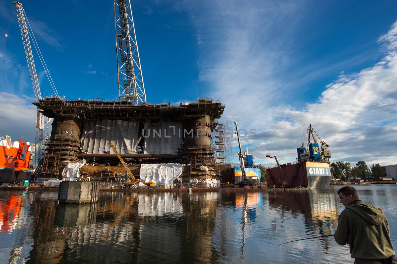 GDANSK, POLAND - SEPTEMBER 19: Unidentified fisher man fishing in front of a docking oil rig at the Gdansk Shipyard under construction with a clear blue sky in the background. The oil rig weights 12 thousand tons. Gdansk, Poland, 2013