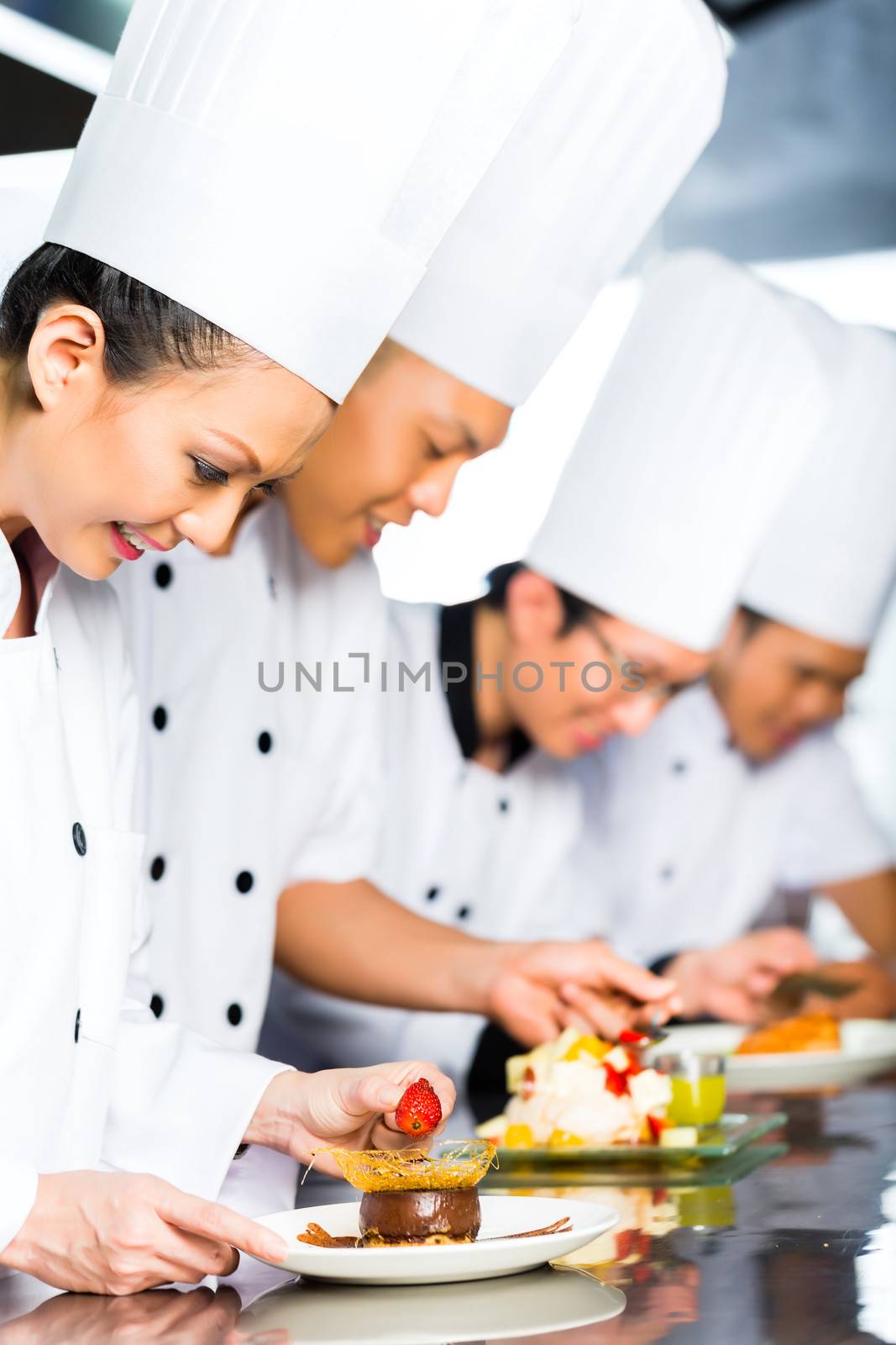 Asian Indonesian chef along with other cooks in restaurant or hotel kitchen cooking, finishing dish or plate for dessert