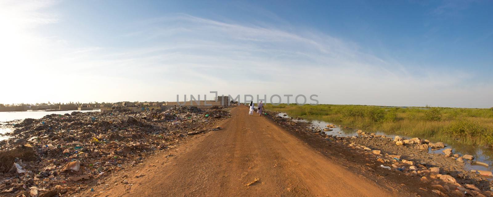 SENEGAL, SAINT-LOUIS, NOVEMBER 30: Two unidentified women walking on an empty road with trashes on the left side and green life on the other site