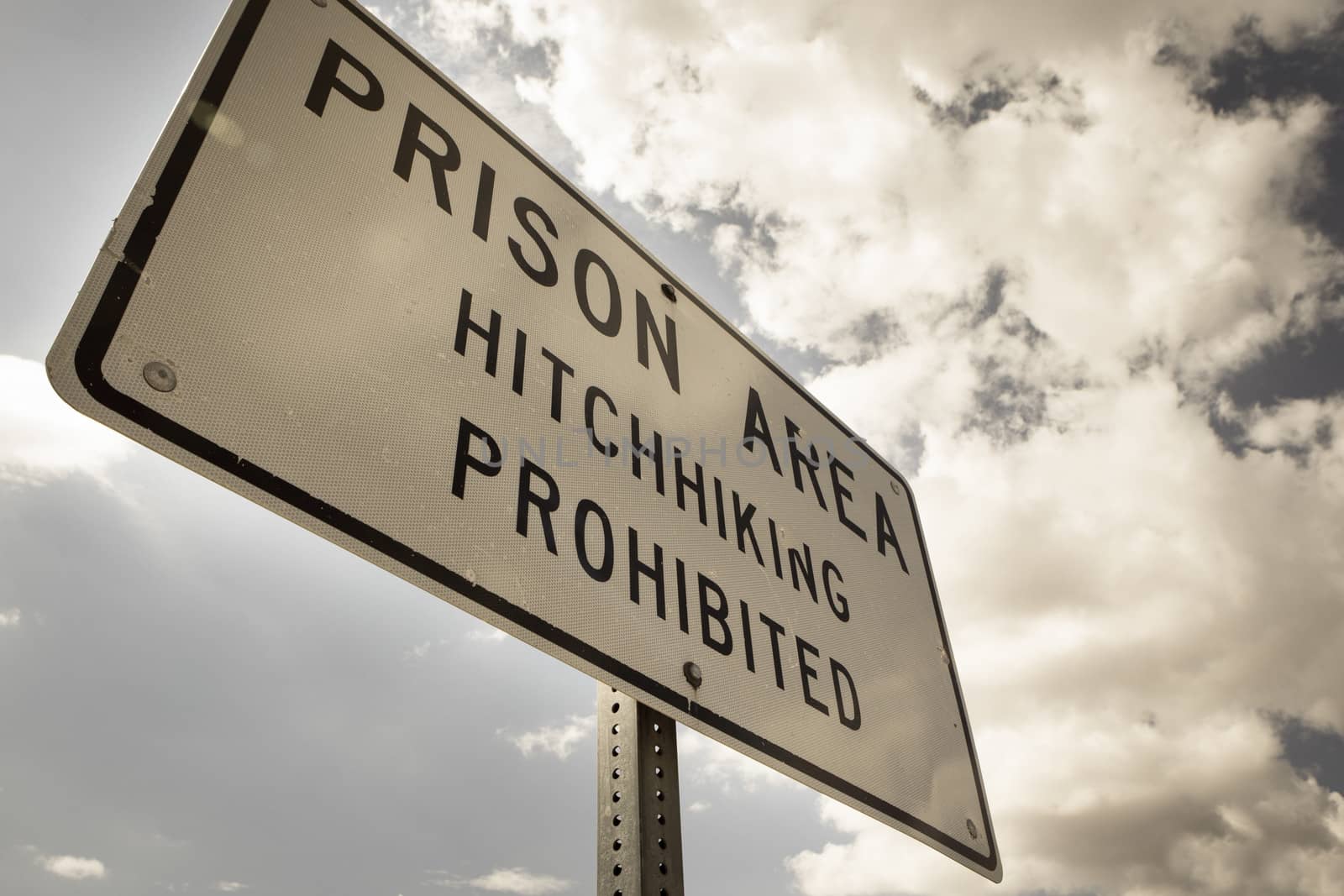 Sign reading "Prison area, hitchhiking prohibited" stands in front of a prison/penitentiary in the desert landscape beneath a cloud filled blue sky. Taken in the Utah state. Retro style image