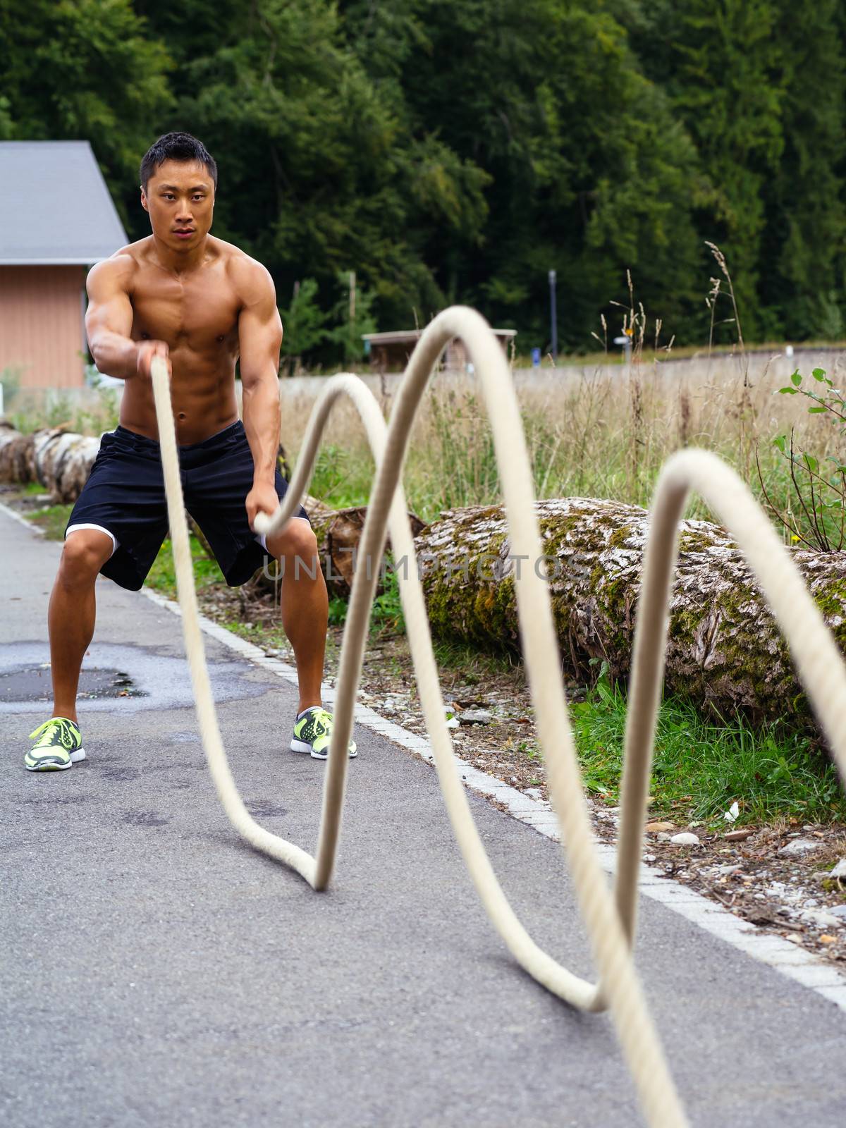 Photo of a muscular Asian man outdoors working out with training ropes.
