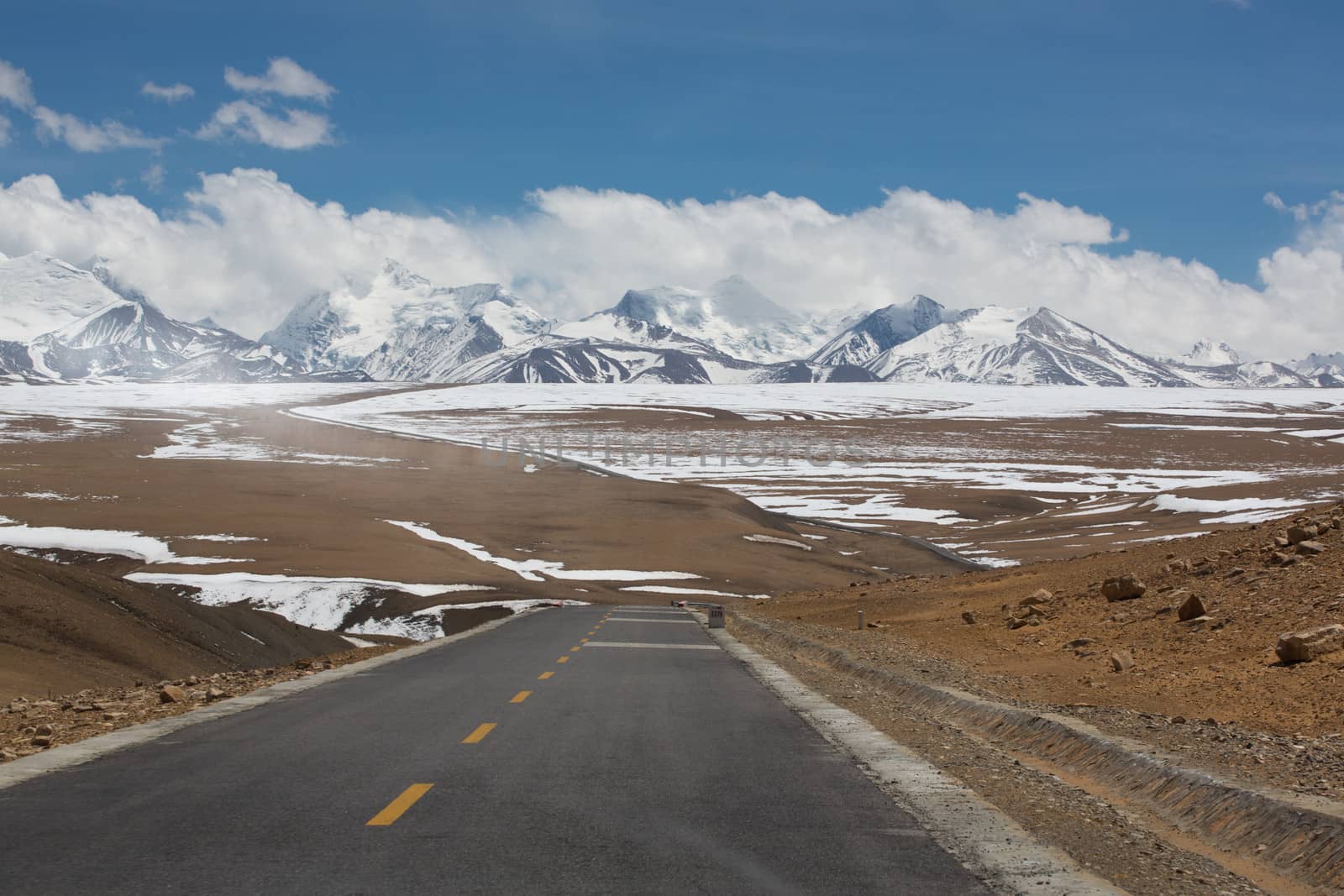 Straight road with a yellow dotted line in the middle with Tibetan landscape of Mountains in the background. The road seems to go toward the top of the snowed mountain in the cloud.