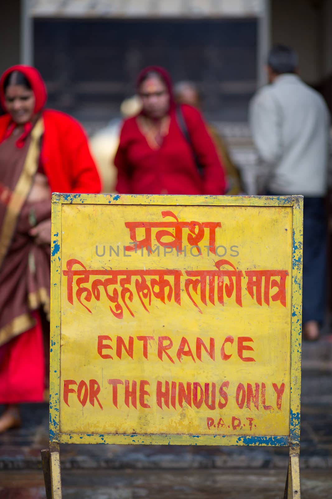 Yellow signboard in front of a Hindu temple in Kathmandu. The message says that the entrance of the temple is for hindus only. In the background, two red unrecognizable woman dressed in red. Nepal 2013.