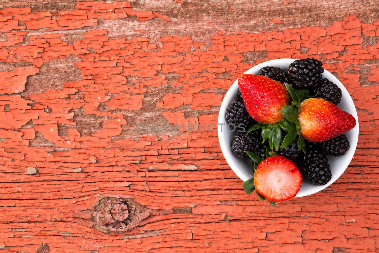 View from above of a small bowl of fresh ripe mixed berries containing strawberries and blackberries on a rustic grungy old wooden picnic table with peeling red paint and copyspace for your text