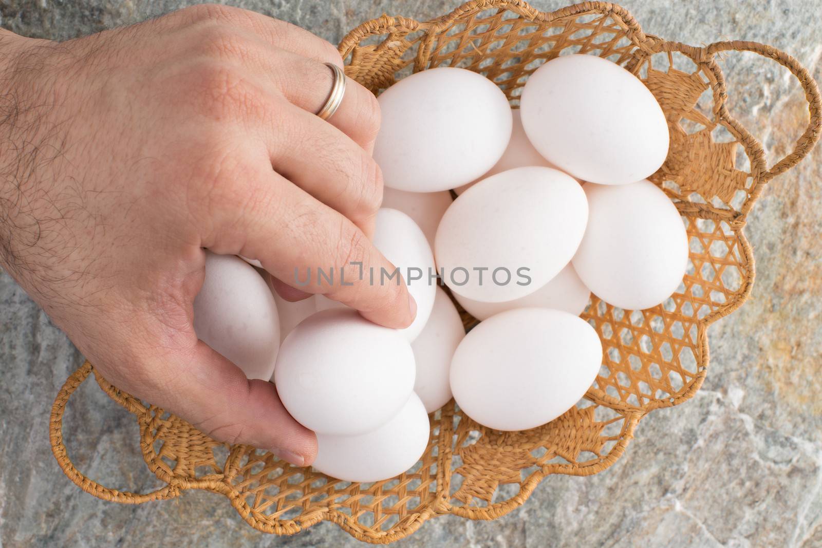 Hand of a man wearing a wedding ring taking a traditional Easter egg from an ornamental woven wicker Easter basket filled with fresh white eggs