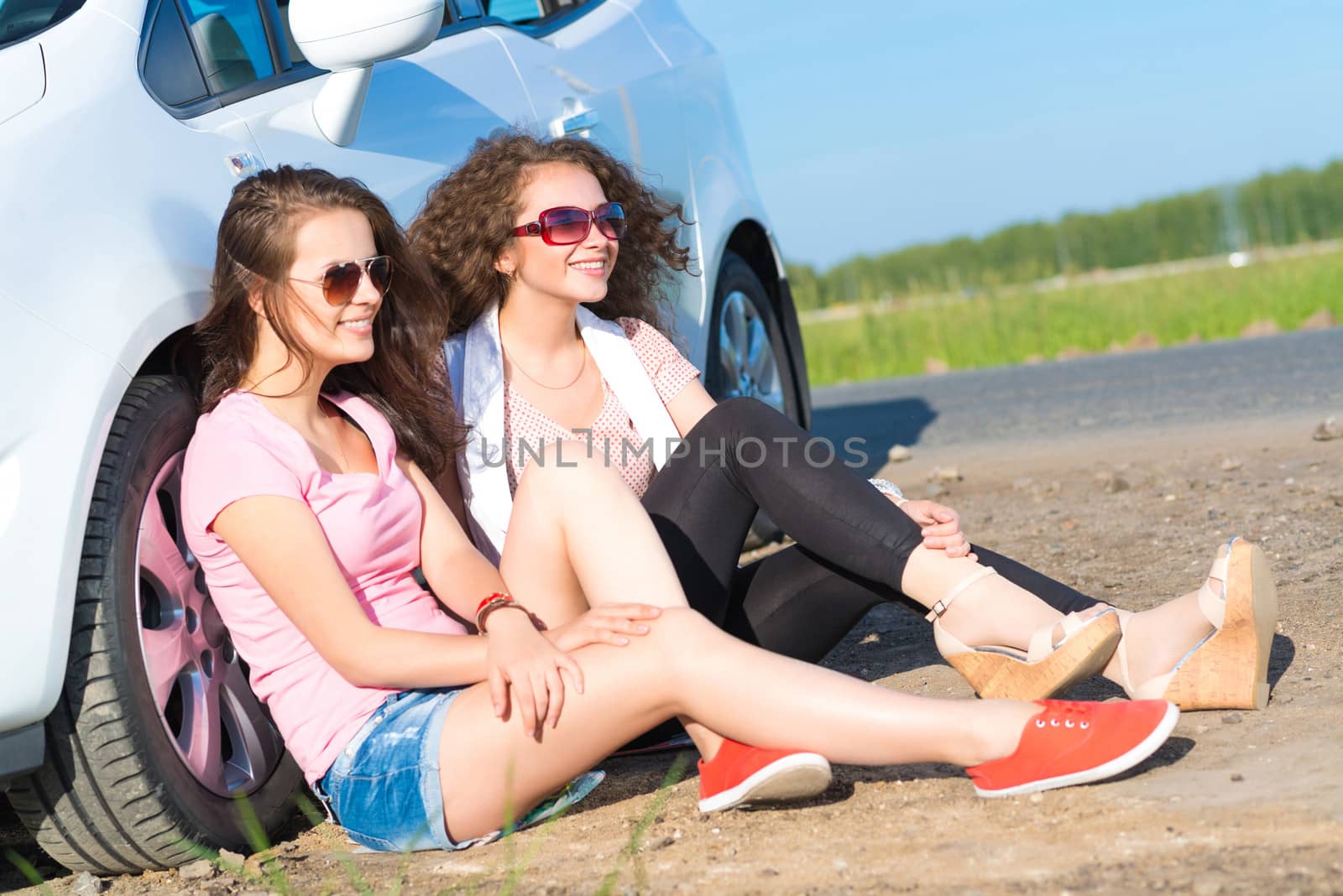 Two attractive young women wearing sunglasses, sitting next to the car