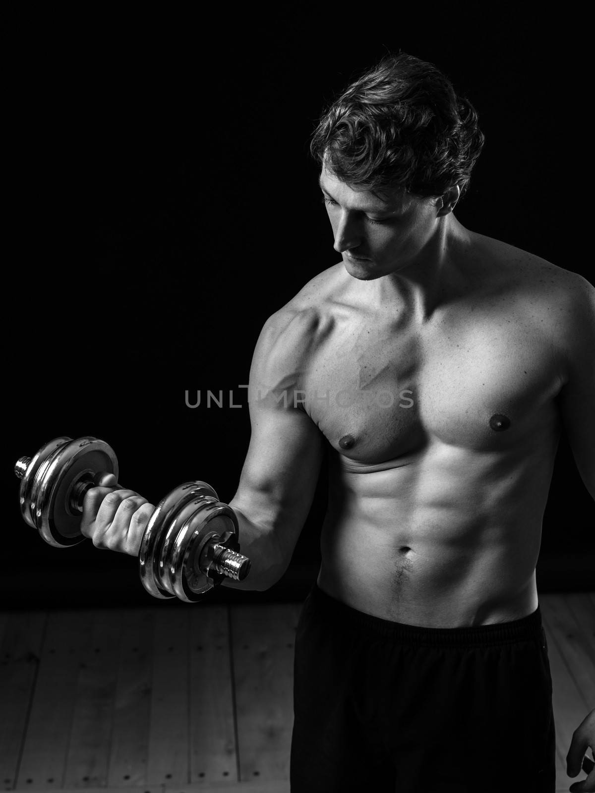 Photo of a man in his early thirties doing bicep curls with a dumbbell over a black background. Black and white version.