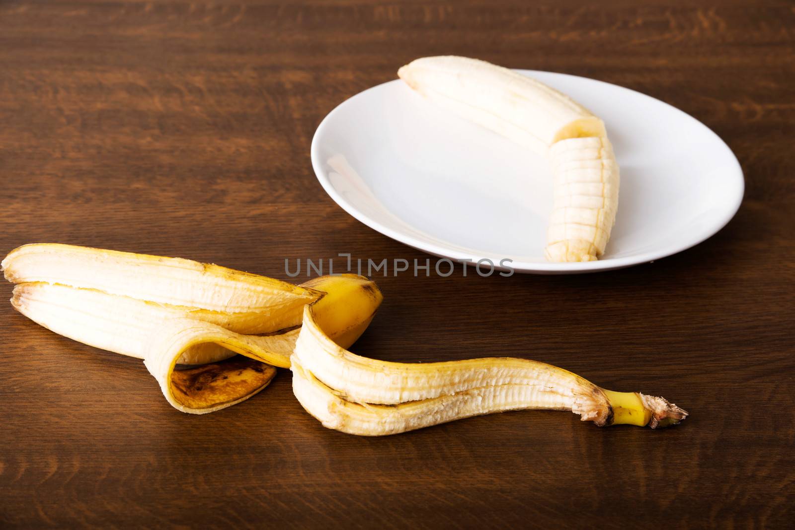 Pilled banana on a plate and its skin lying next to it. Over wooden background.