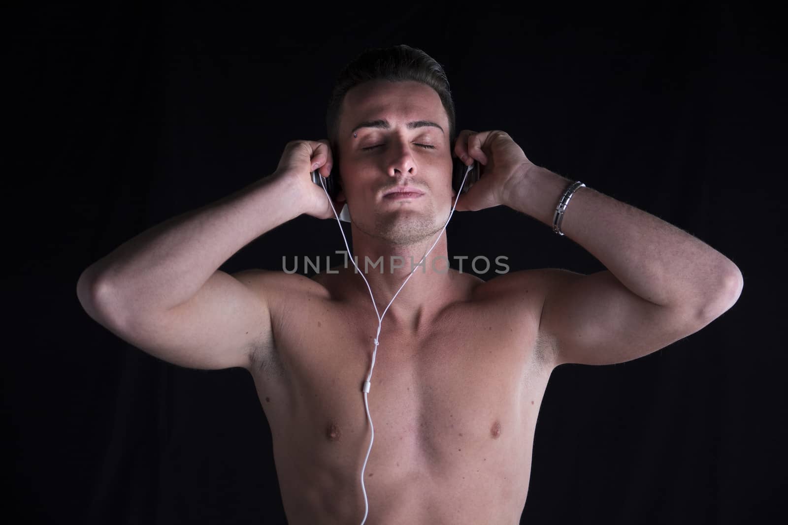 Athletic shirtless man listening to music on headphones, eyes closed, black background