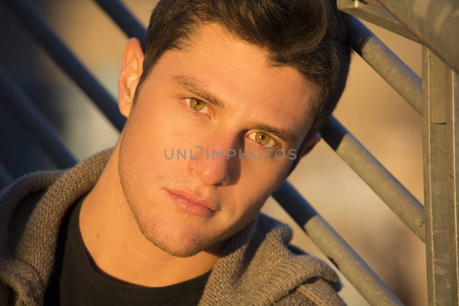 Headshot of handsome young man, lit by sunset light, leaning against handrail