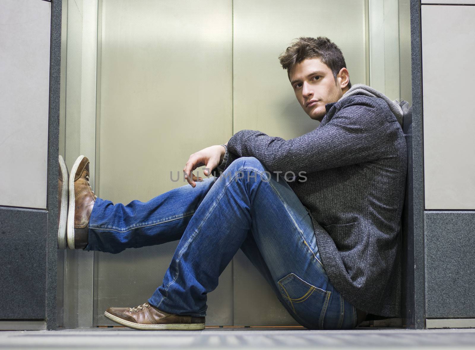 Handsome young man sitting in front of elevator (lift) doors, looking at camera