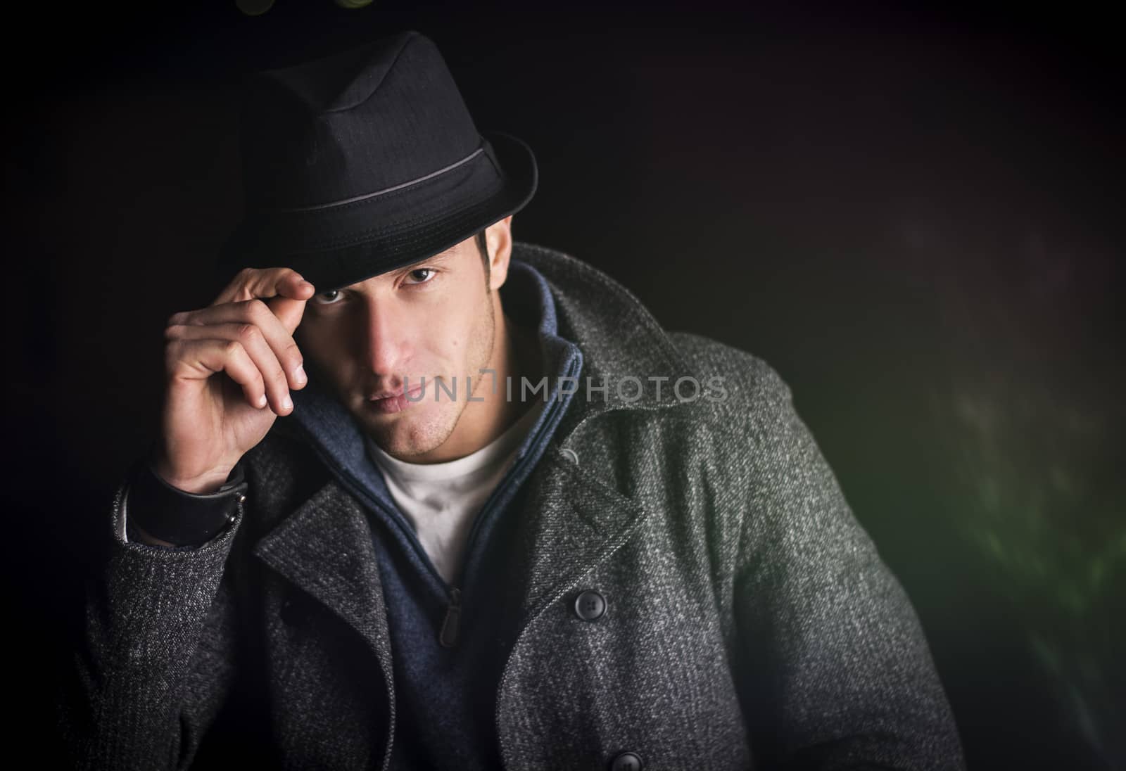 Attractive young man at night, wearing winter coat, tipping fedora hat while looking at camera