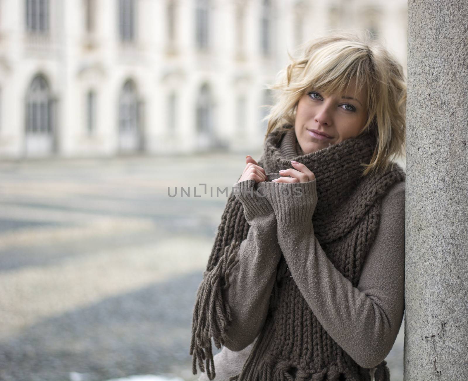 Pretty young blonde woman outdoors, wearing wool scarf and sweater, leaning on marble column