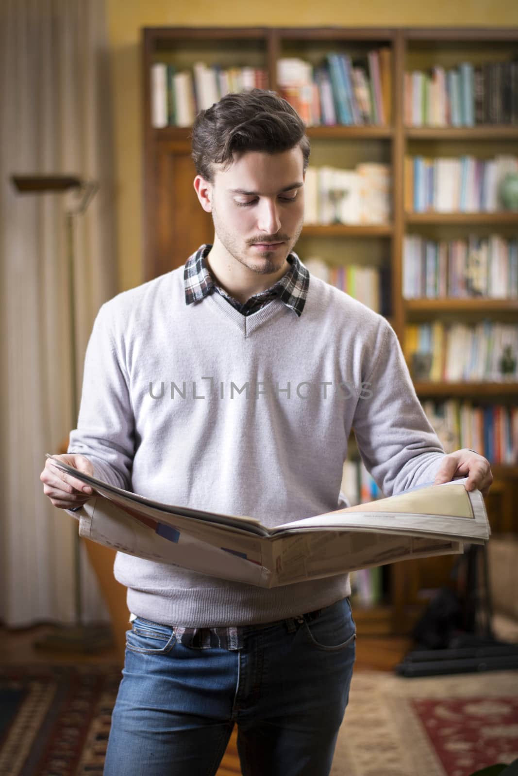 Handsome young man reading newspaper at home in his living-room, standing