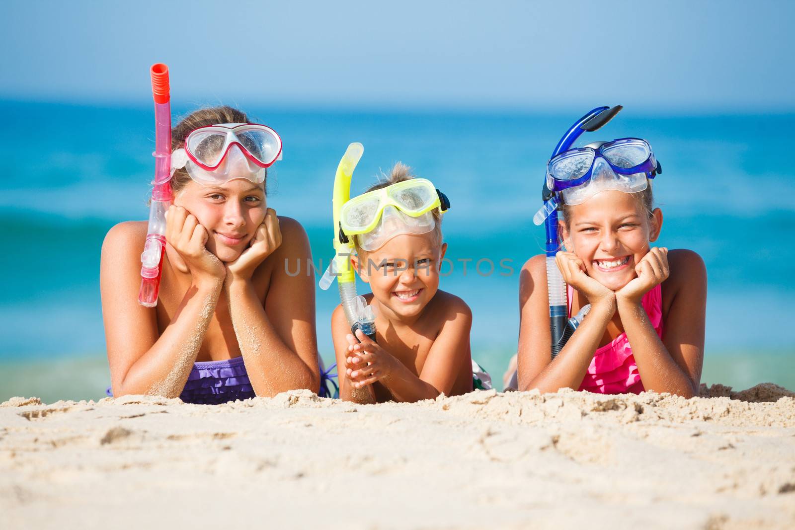 Three happy children on beach with colorful face masks and snorkels, sea in background.