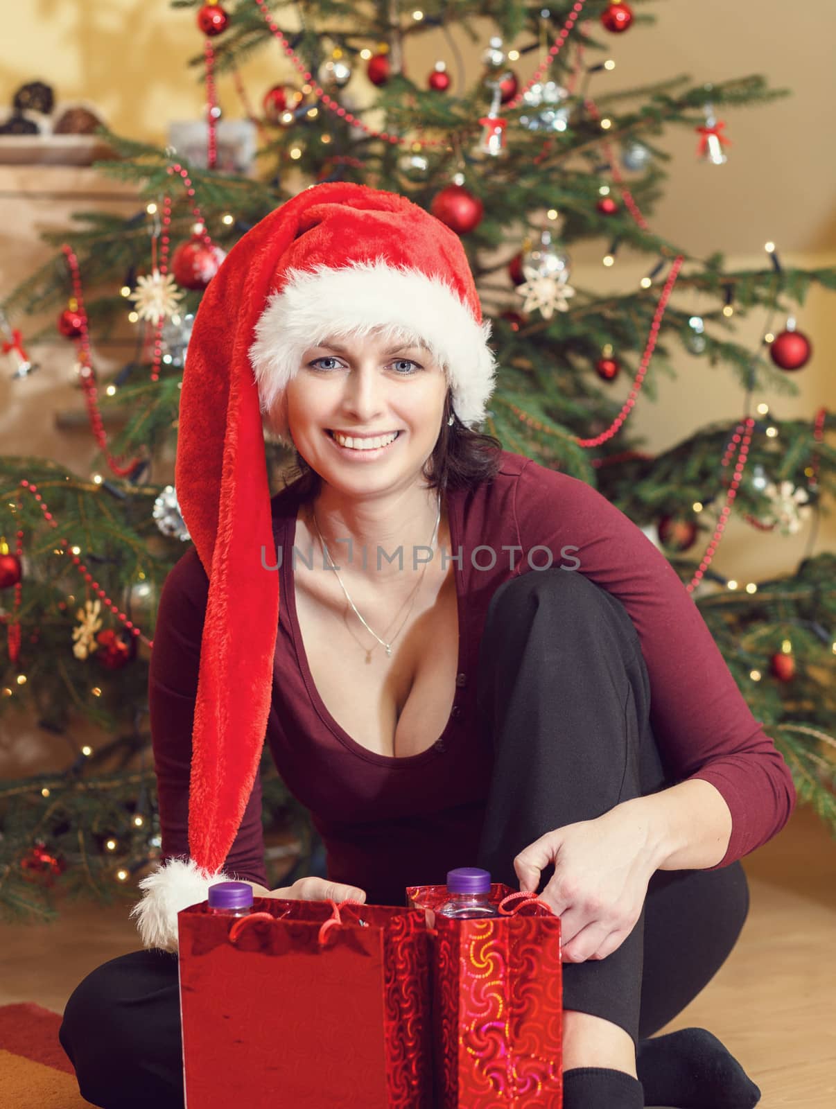 Beautiful middle age woman in red party santa hat sitting near the Christmas tree with gifts