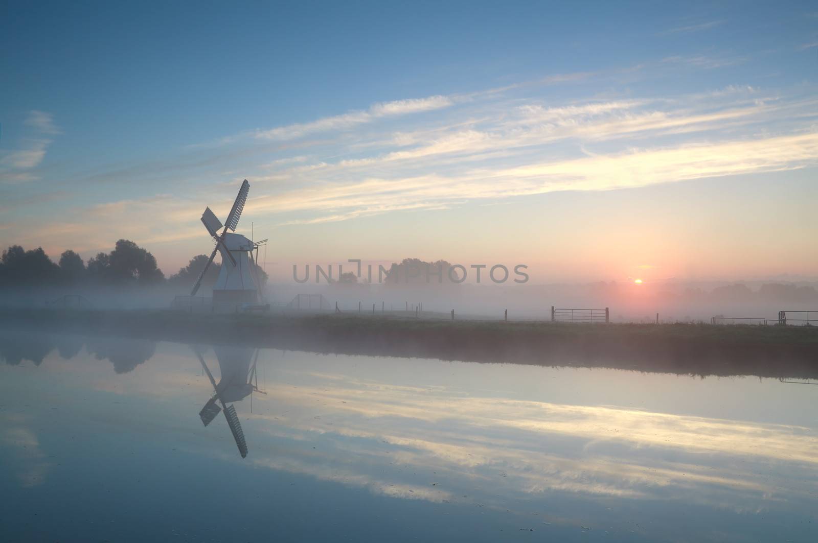 Dutch windmill by river during misty sunrise by catolla