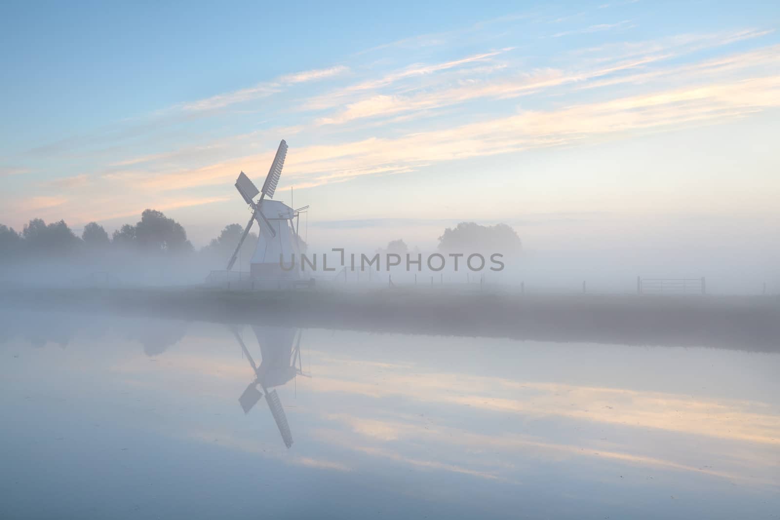 Dutch windmill in dense fog during sunrise by river, Holland
