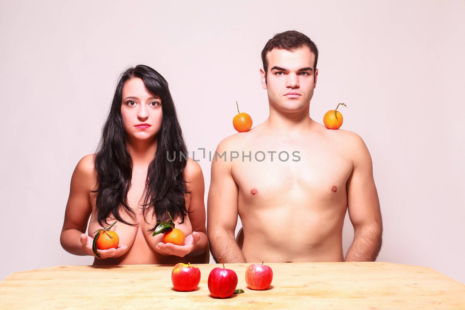 Conceptual image of a young nude man and woman posing with fresh tropical fruit in their hands and balanced on their shoulders looking at the camera with serious expressions as they sit at a table