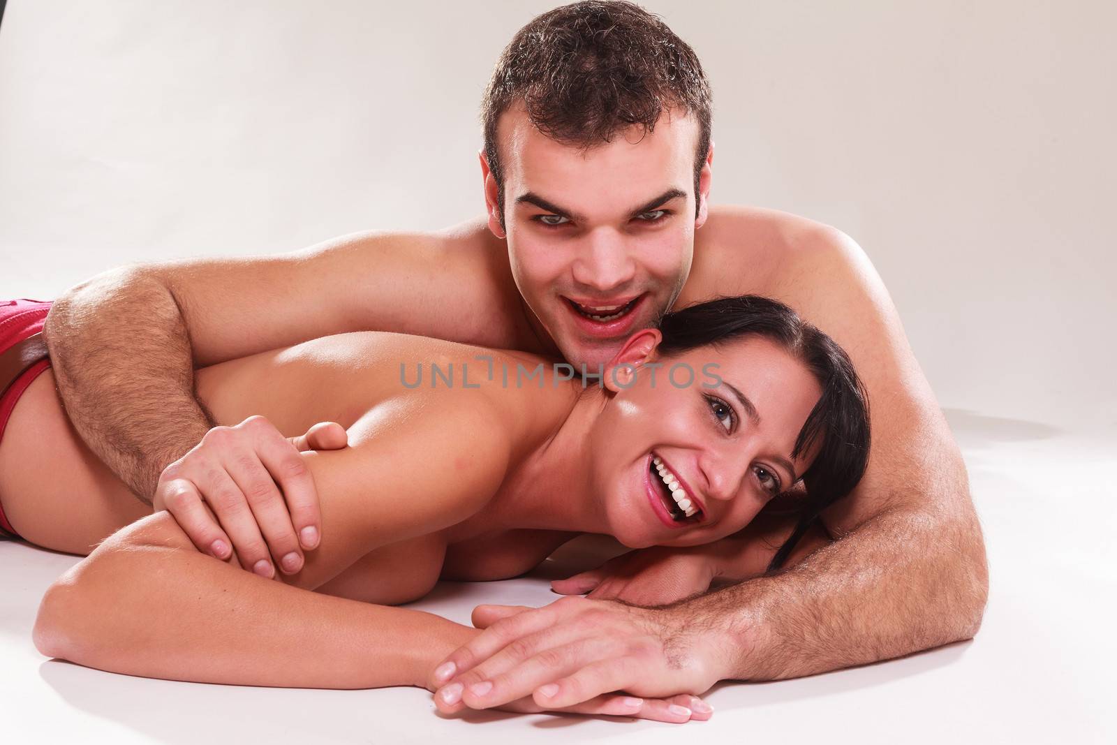 Playful romantic man and woman lying naked on their stomachs in each others arms laughing at the camera,