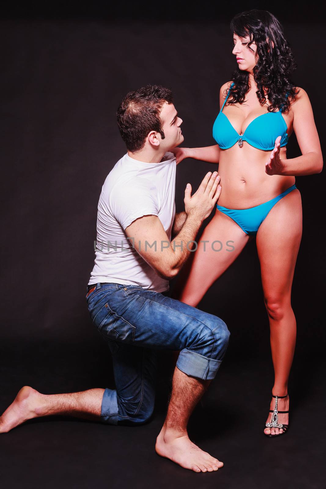 Young couple acting out sexual fantasies of dominance and subservience with the barefoot man kneeling at the womans feet as she stands in her lingerie gripping him by the shirt
