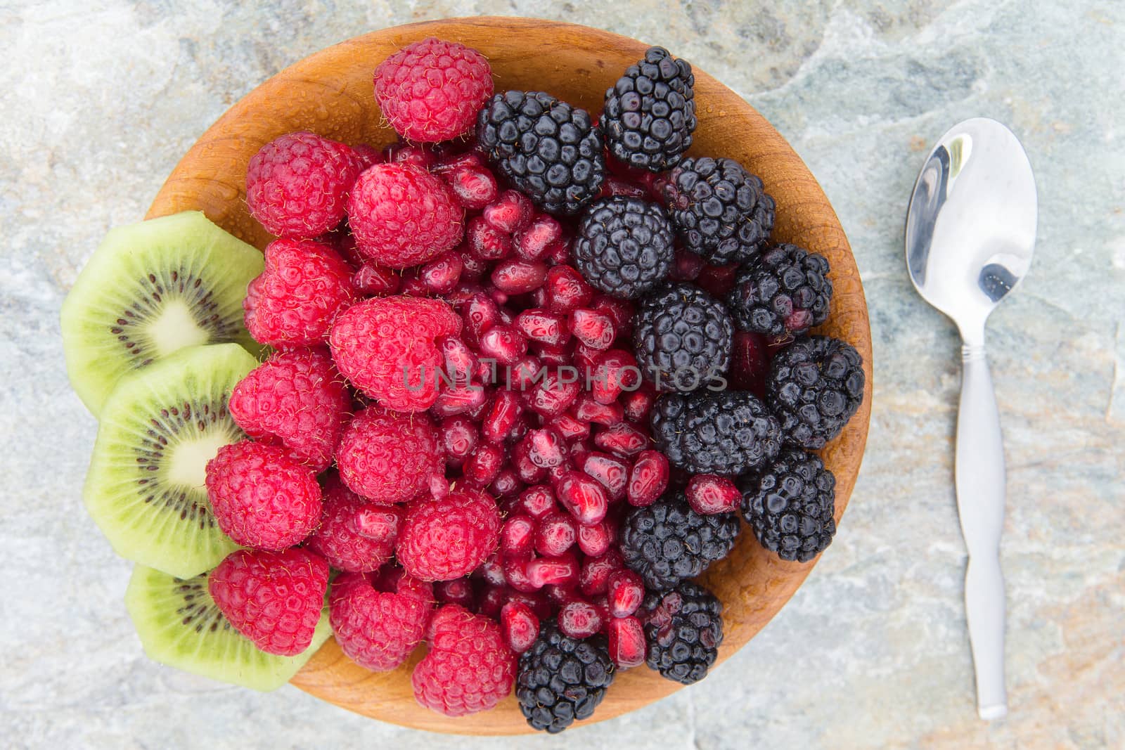 Delicious nutritional snack made of fresh slices of kiwi, seeds of pomegranate, blackberries and raspberries, in a wooden bowl, next to a metallic spoon, on a table, high-angle shot