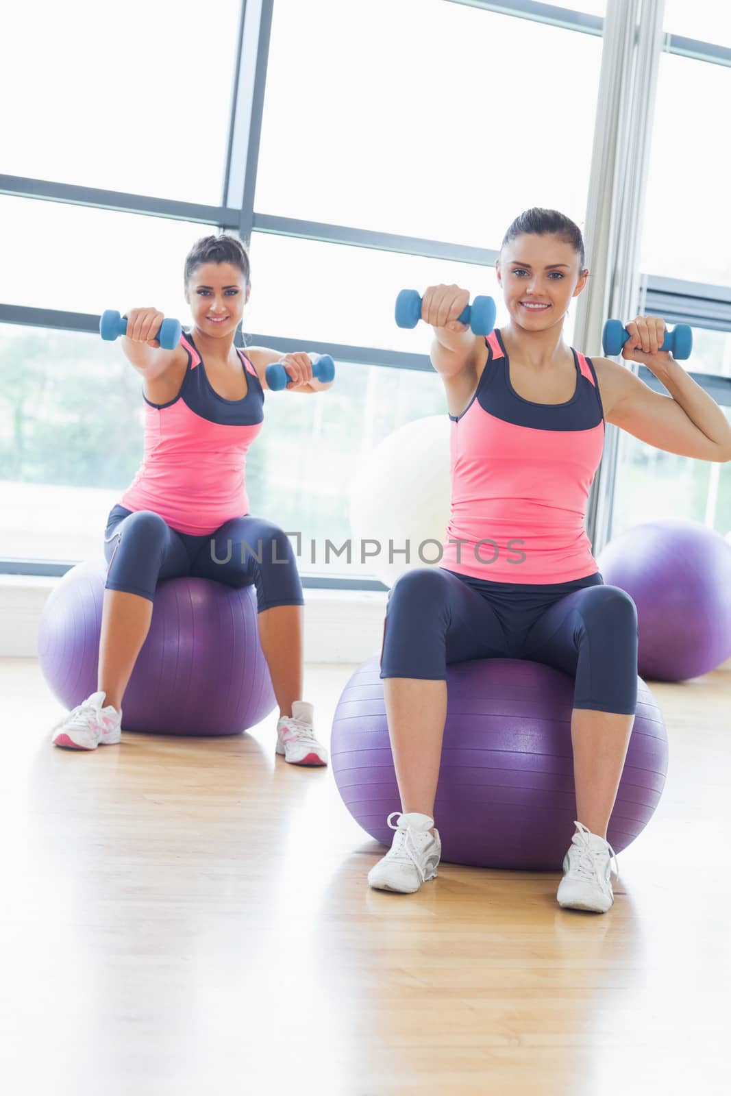 Two fit young women exercising with dumbbells on fitness balls in the bright gym