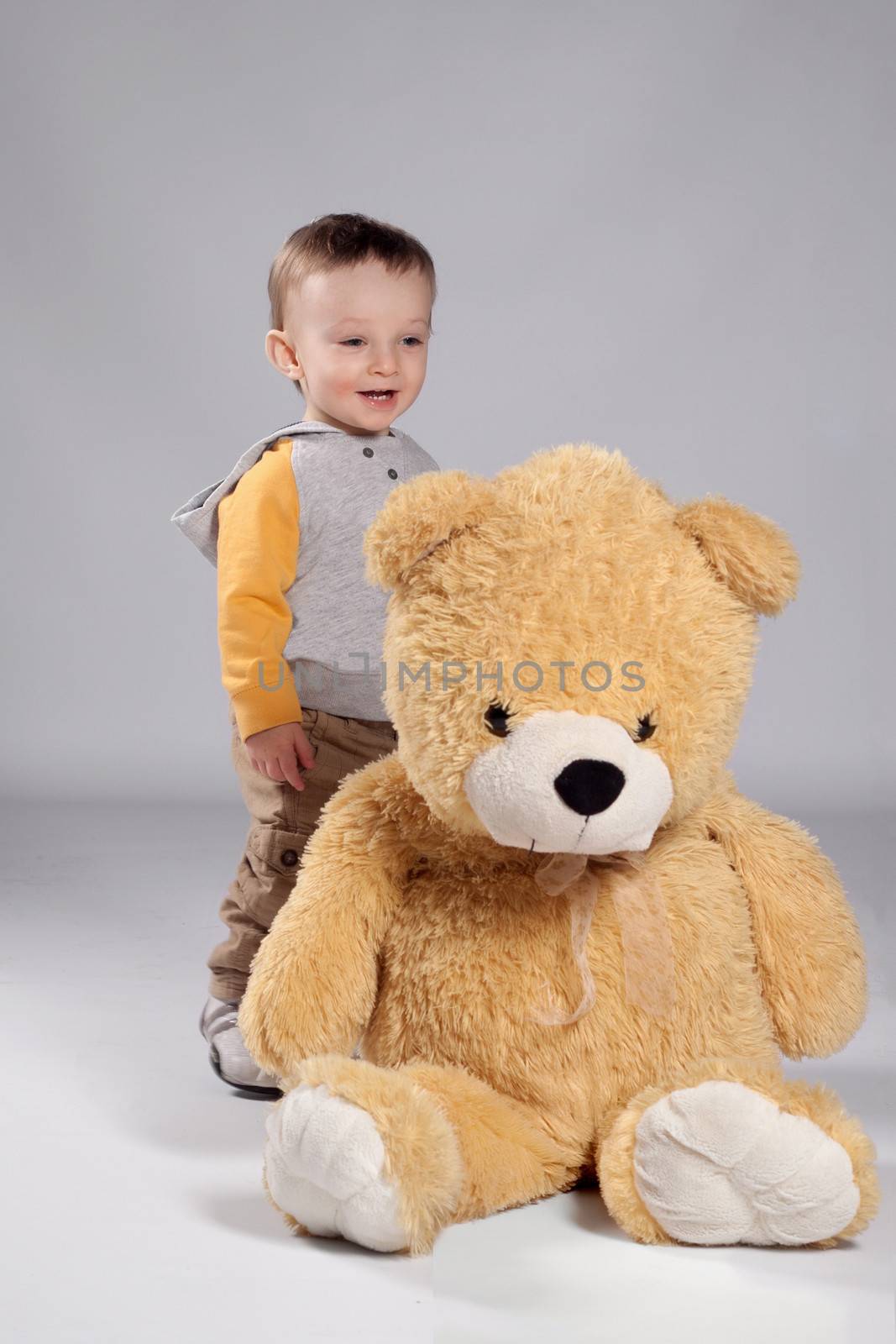 Toddler boy standing in a sweatshirt and sneakers behind a big teddy bear