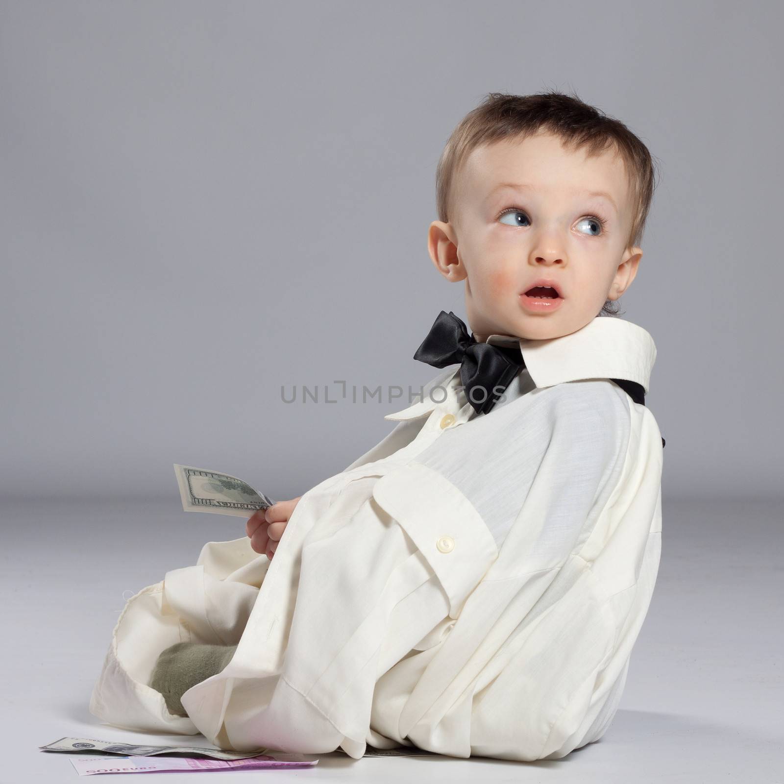 Boy toddler businessman, sitting next to a pile of money dressed in grown-shirt with bow tie, holding in hand the dollars