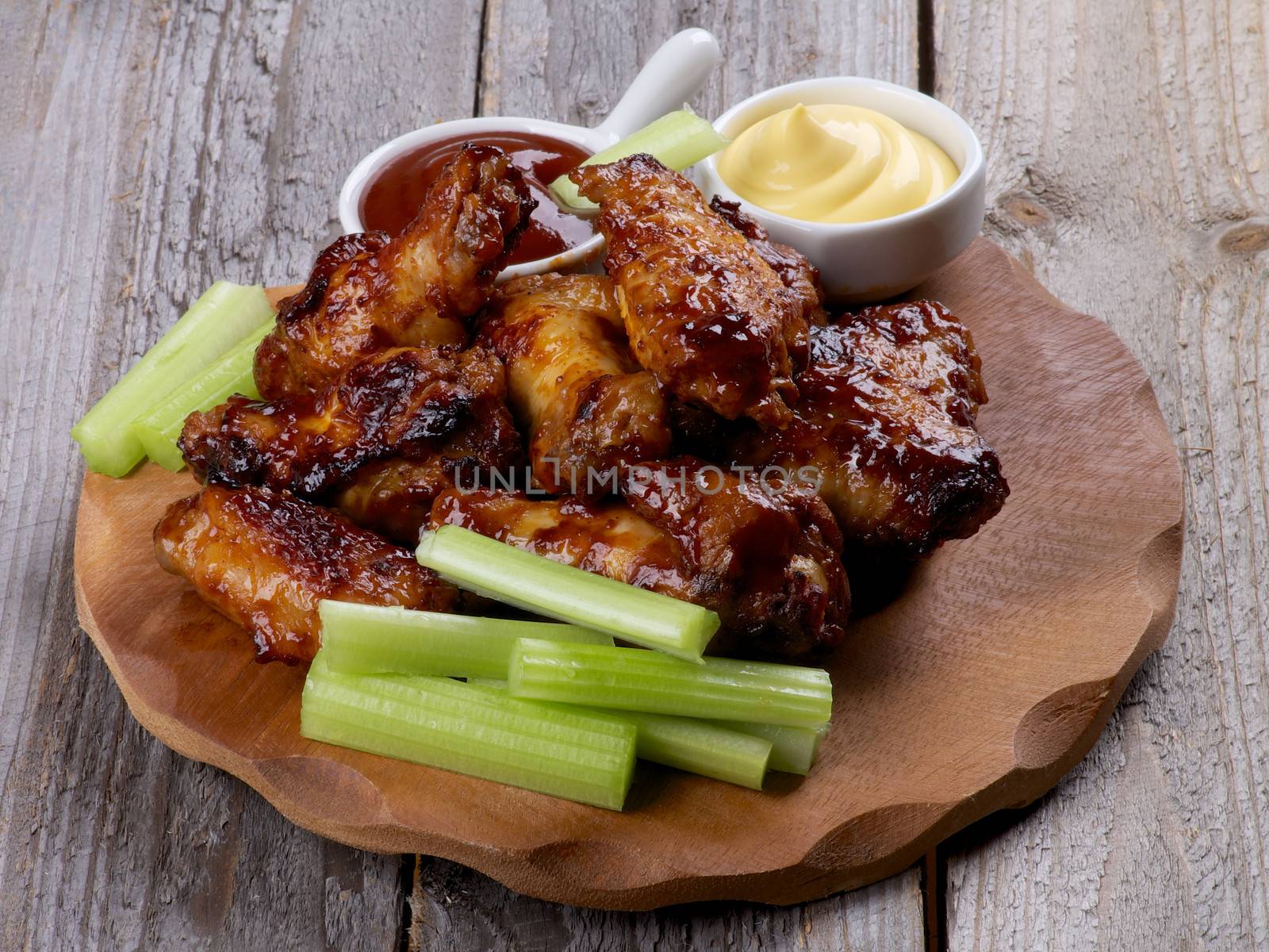 Chicken Legs and Wings Barbecue with Ketchup and Cheese Sauces and Celery Sticks on Wooden Plate closeup