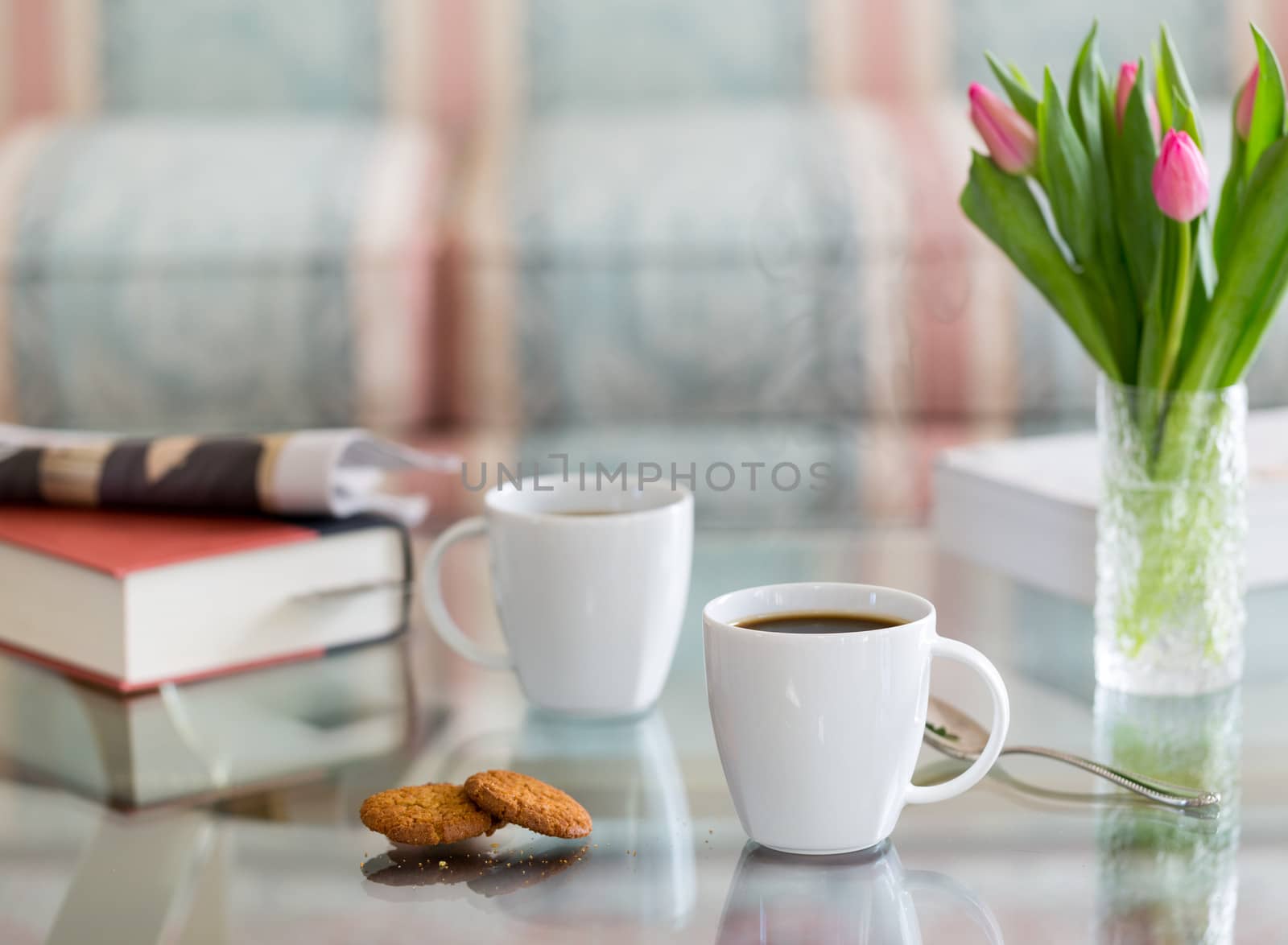 Modern white porcelain cup of black coffee on glass table with spoon and ginger biscuits. Book newspaper and vase of tulips in the background out of focus