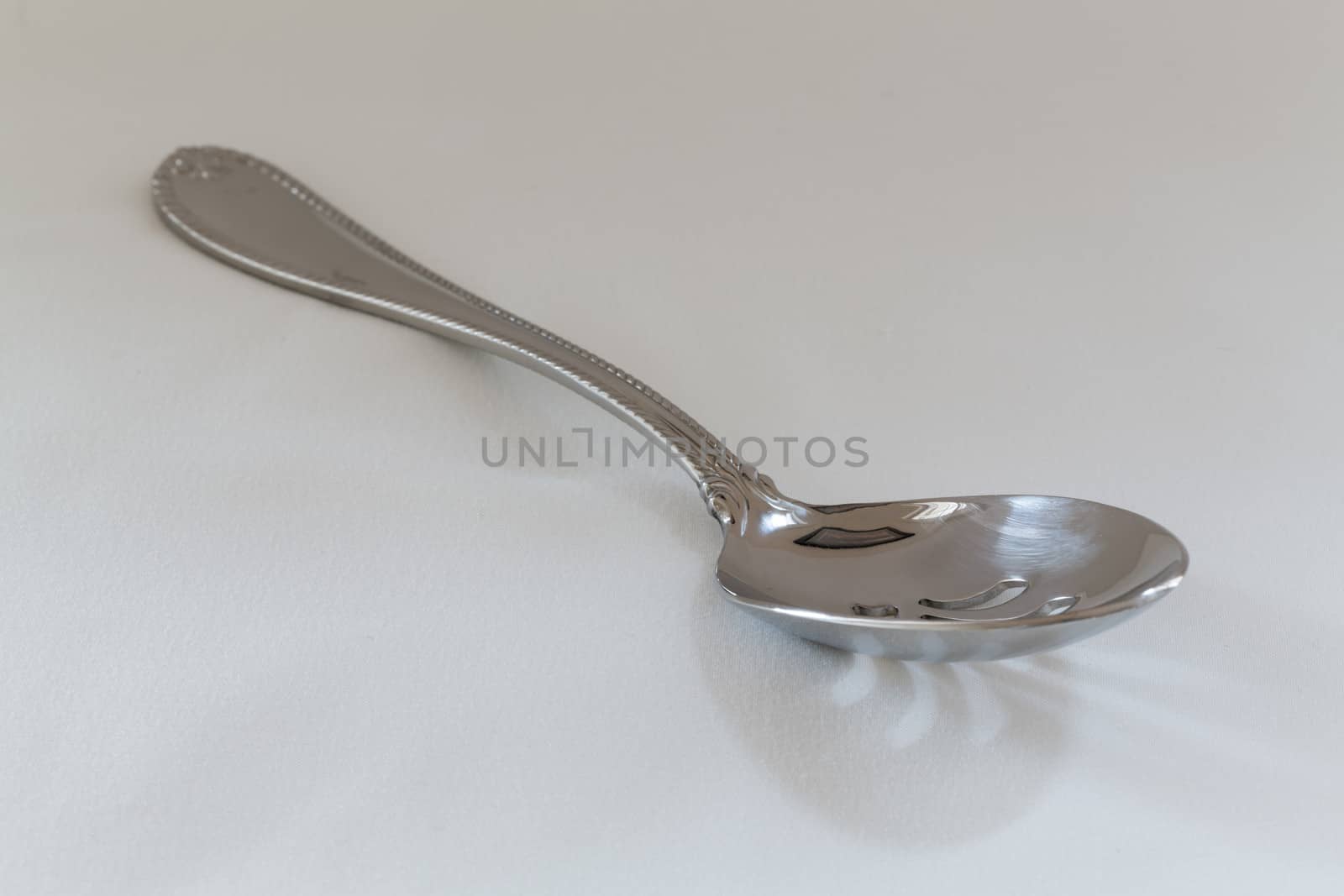 Decorated slotted serving or dessert spoon for draining wet vegetables in side view on fine white tablecloth with pattern just visible