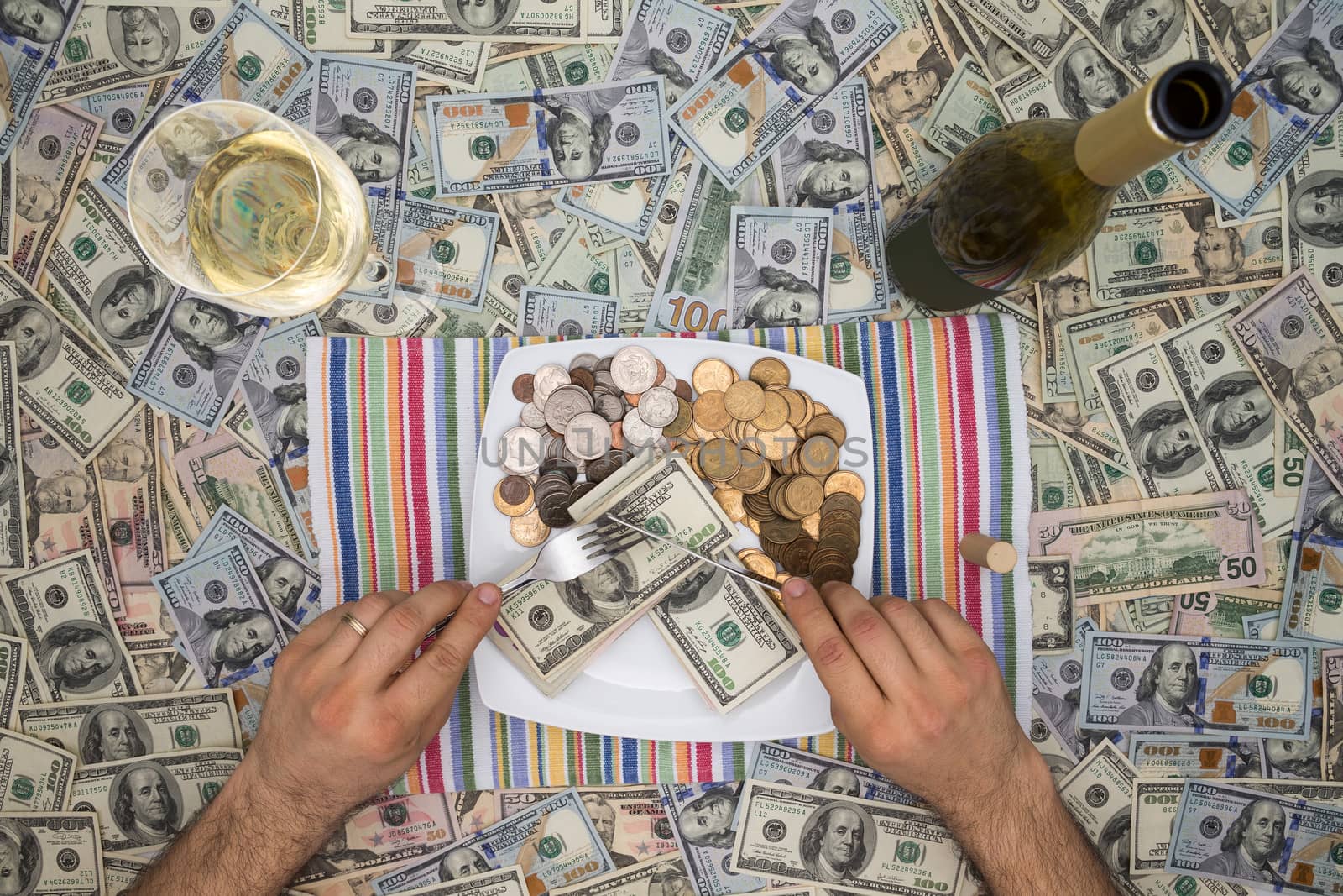 Conceptual image of a man eating money through extravagance with an overhead view of him sitting at a table covered in 100 dollar bills eating money on a plate with a glass and bottle of champagne