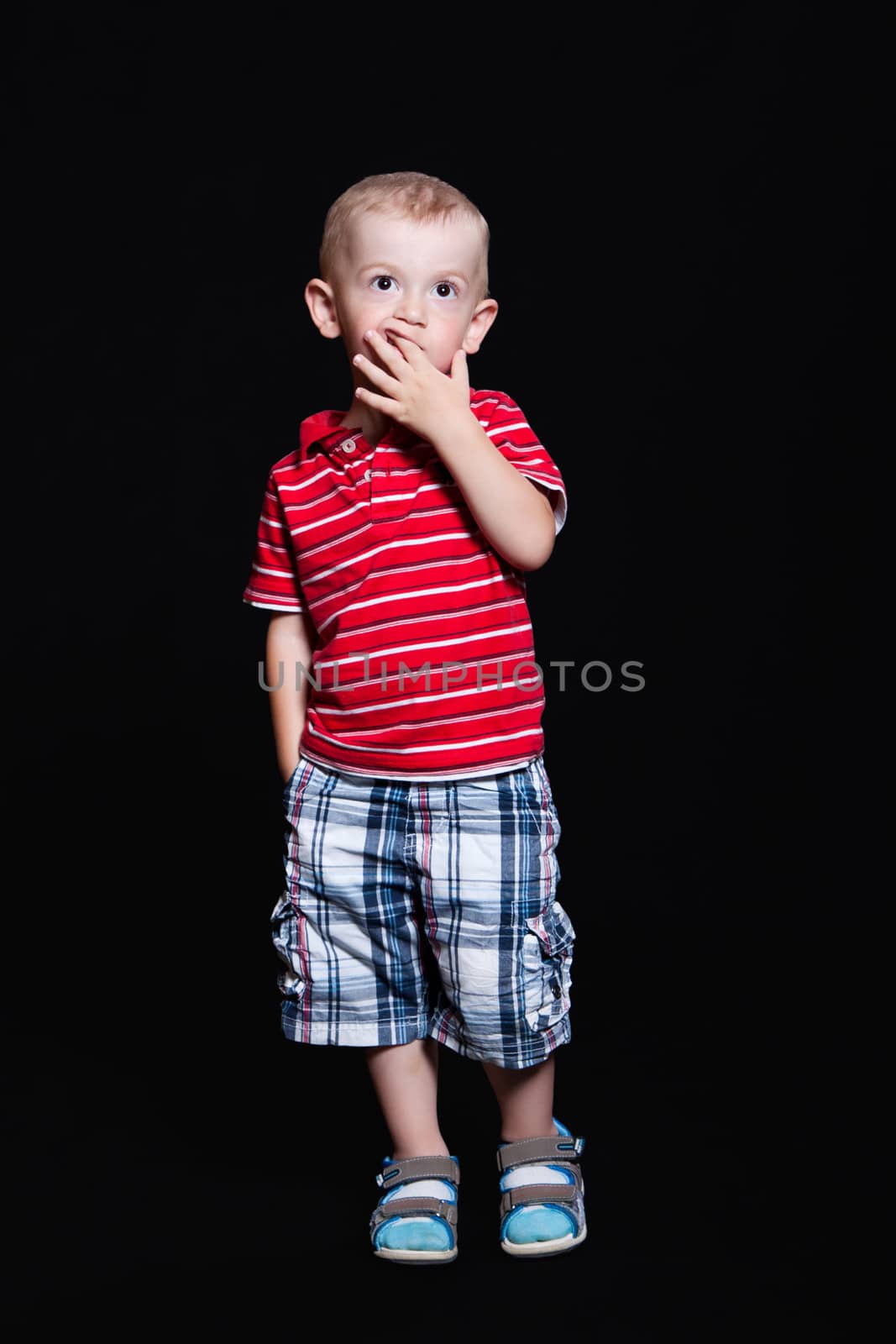 Little boy in a striped shirt and checkered shorts standing with hand in his pocket and hand in mouth