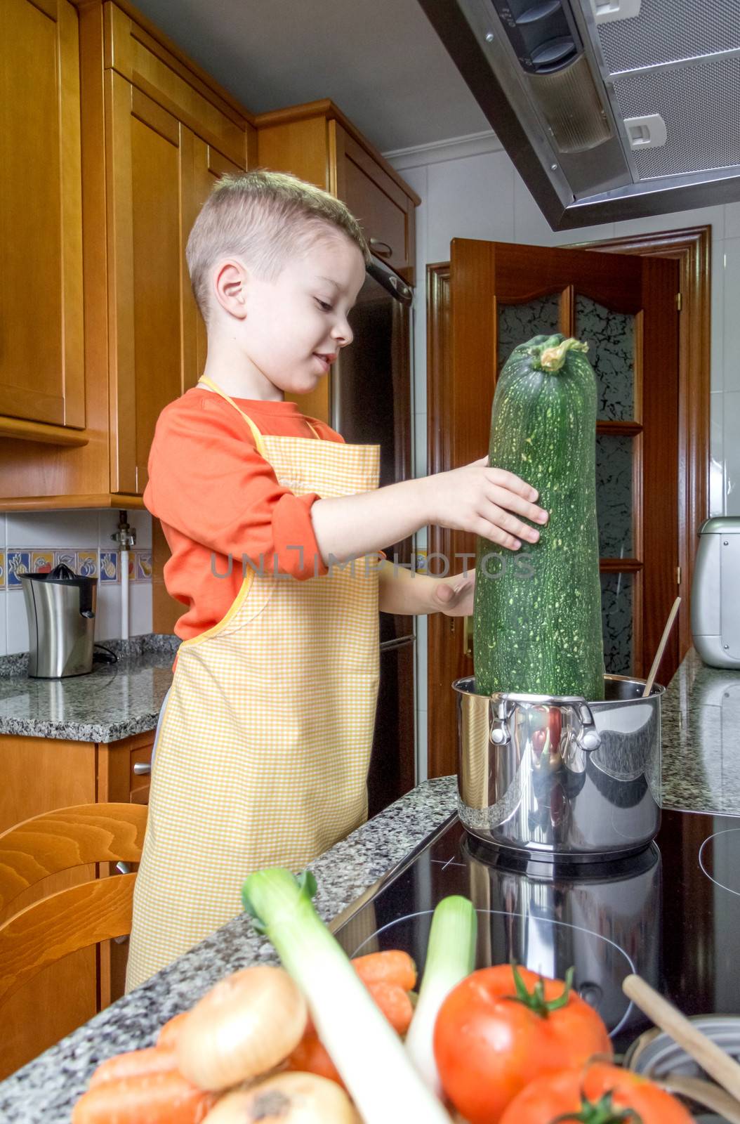 Cute child chef with apron cooking big zucchini and other vegetables in a pot on the kitchen