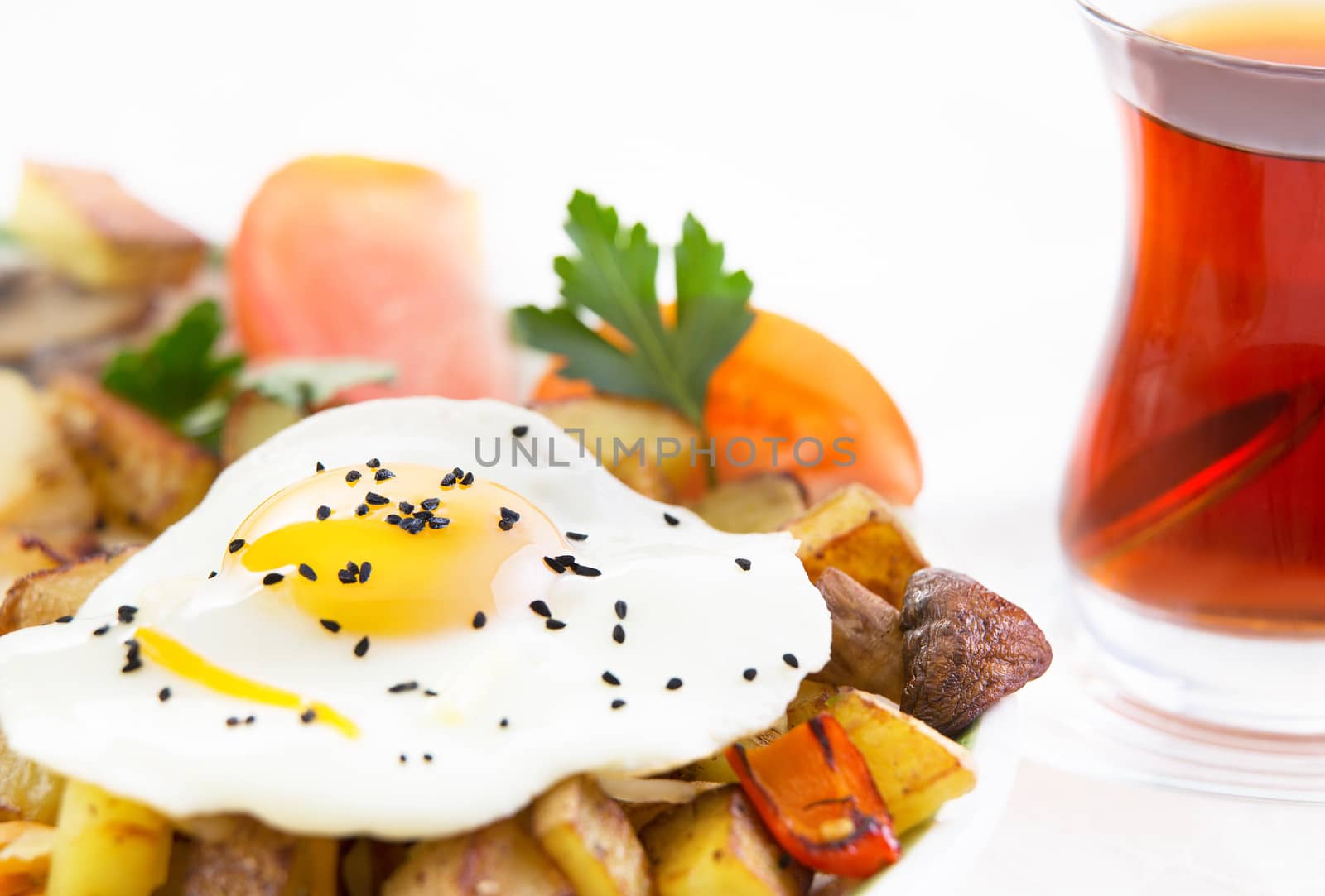 Sunny side up fried egg served on potatoes garnished with mushrooms and sliced fresh tomato accompanied by a glass of hot black Turkish tea on a white background