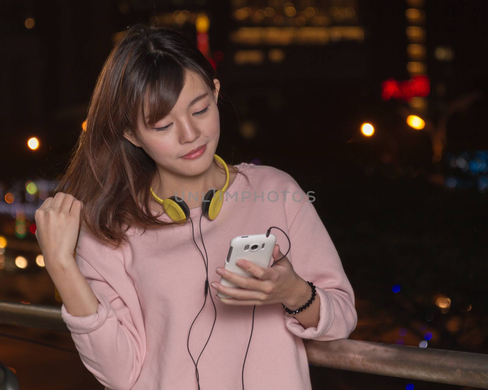 Attractive Malaysian female wearing headphones and holding a cell phone with city lights in background