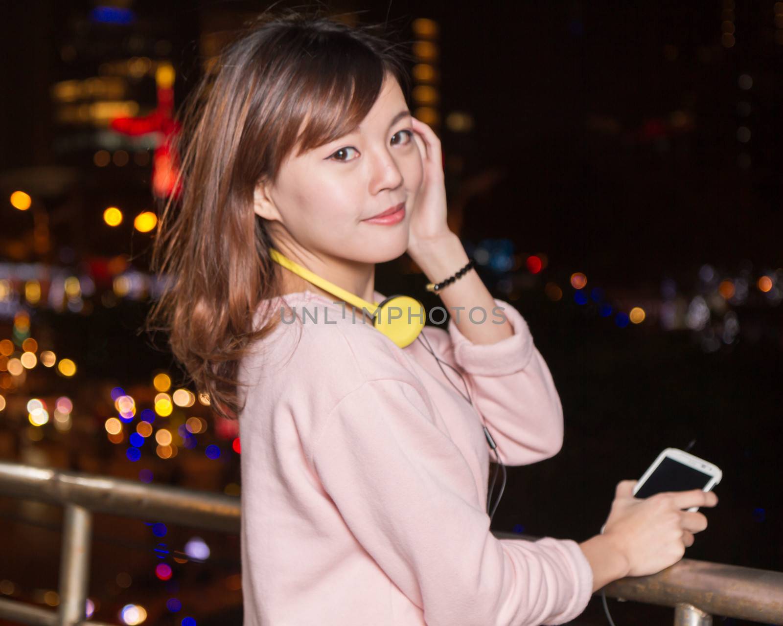 Attractive Malaysian female wearing headphones and holding a cell phone with city lights in background