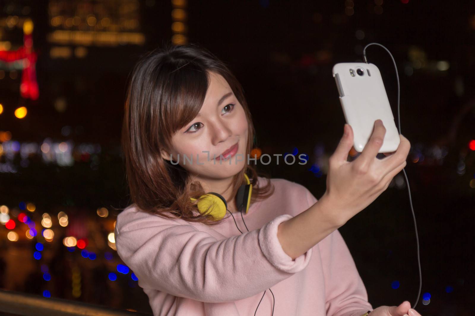 Attractive Malaysian female wearing headphones and holding a cell phone with city lights in background