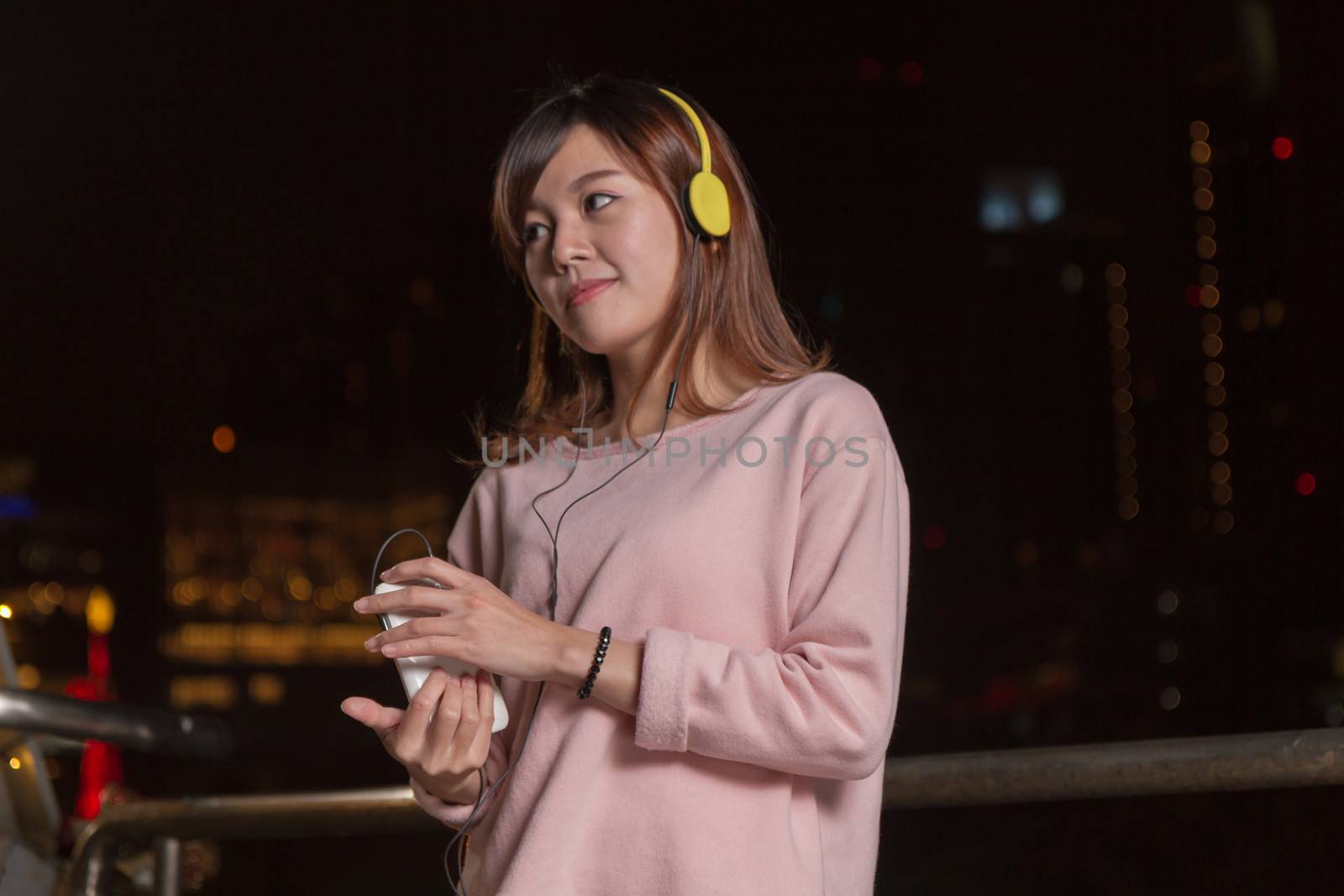 Attractive Malaysian female wearing headphones and holding a cell phone with city lights in background