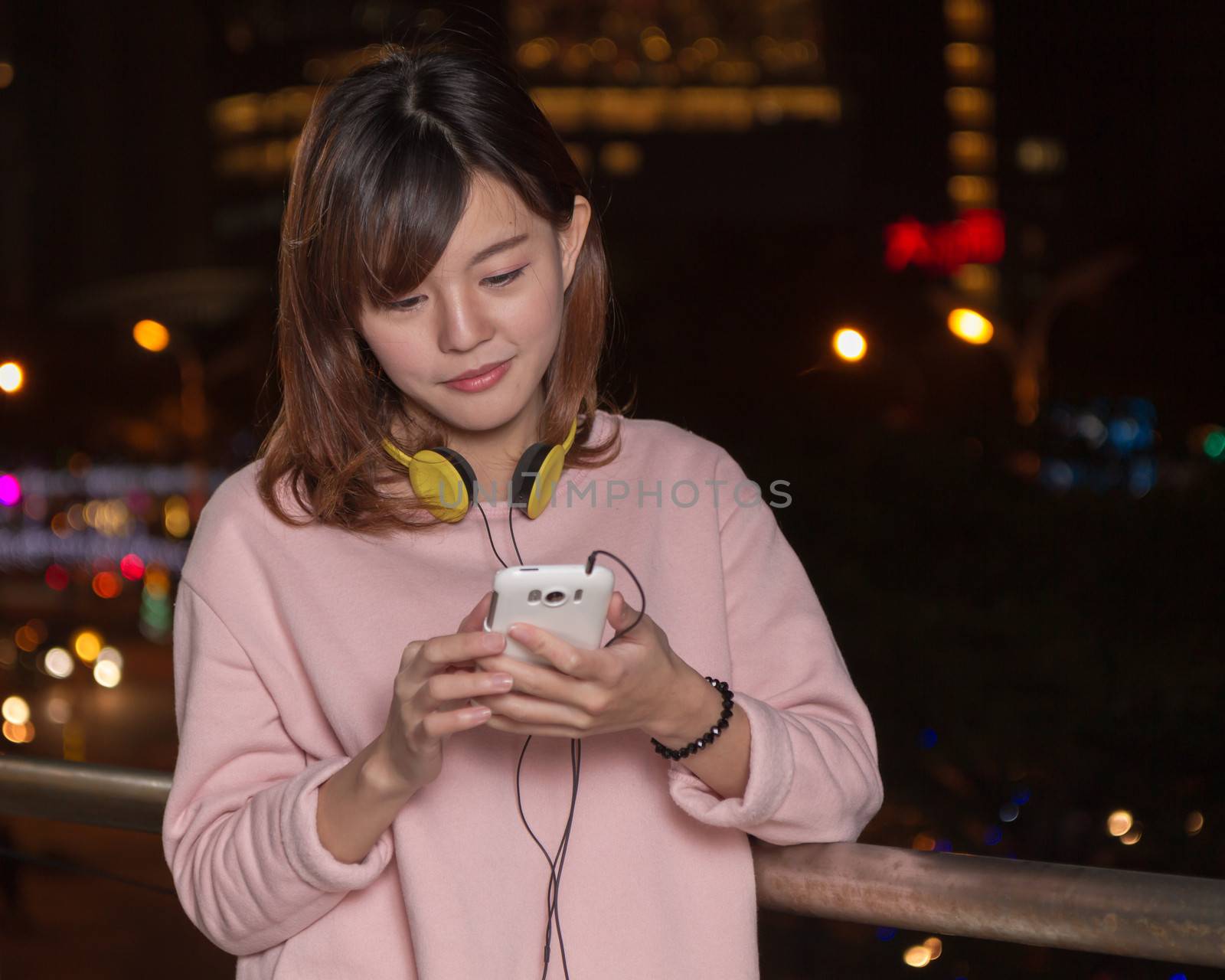 Attractive Malaysian female wearing headphones and holding a cell phone with city lights in background