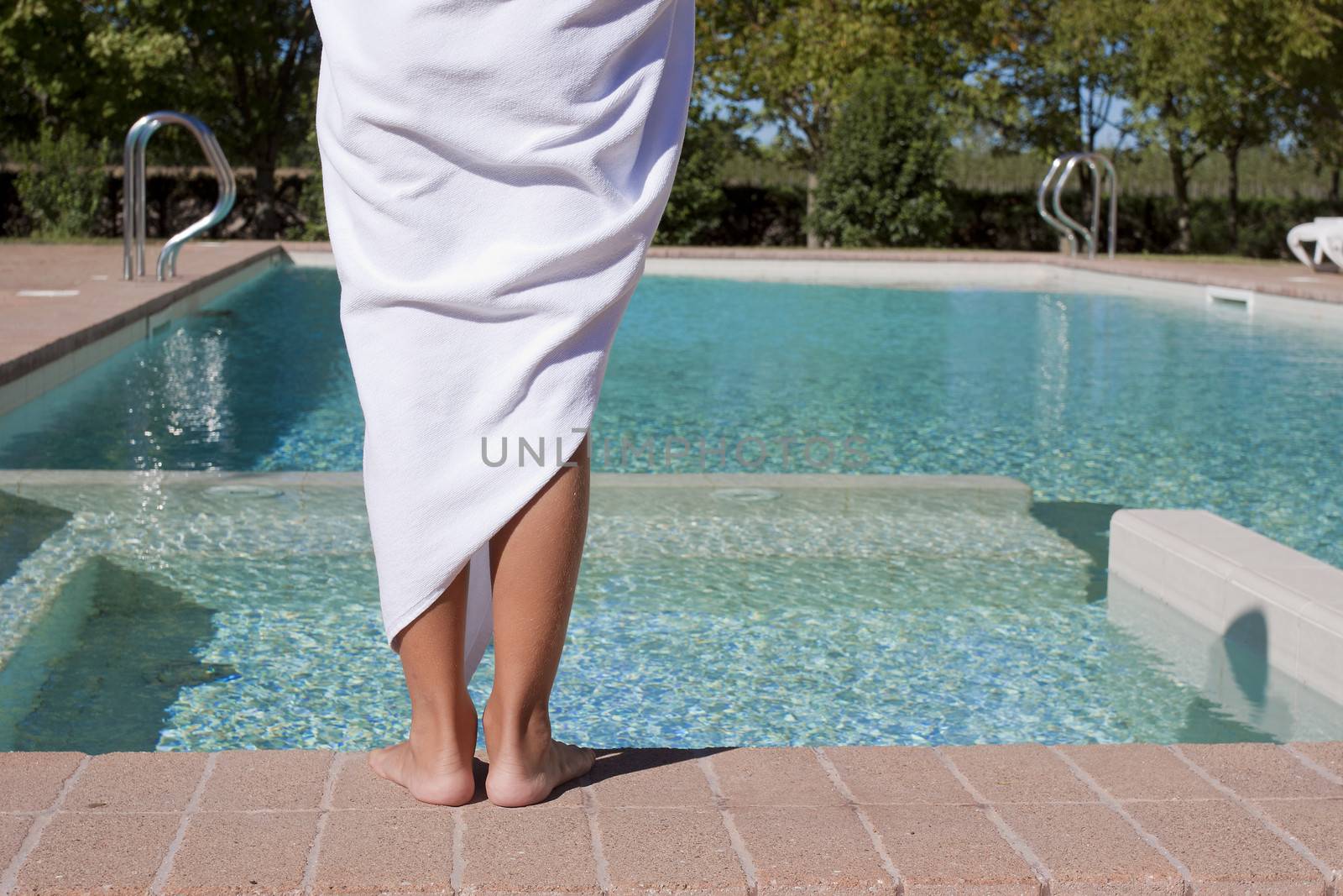 A person wrapped up in a towel standing with back to camera looking at an empty swimming pool