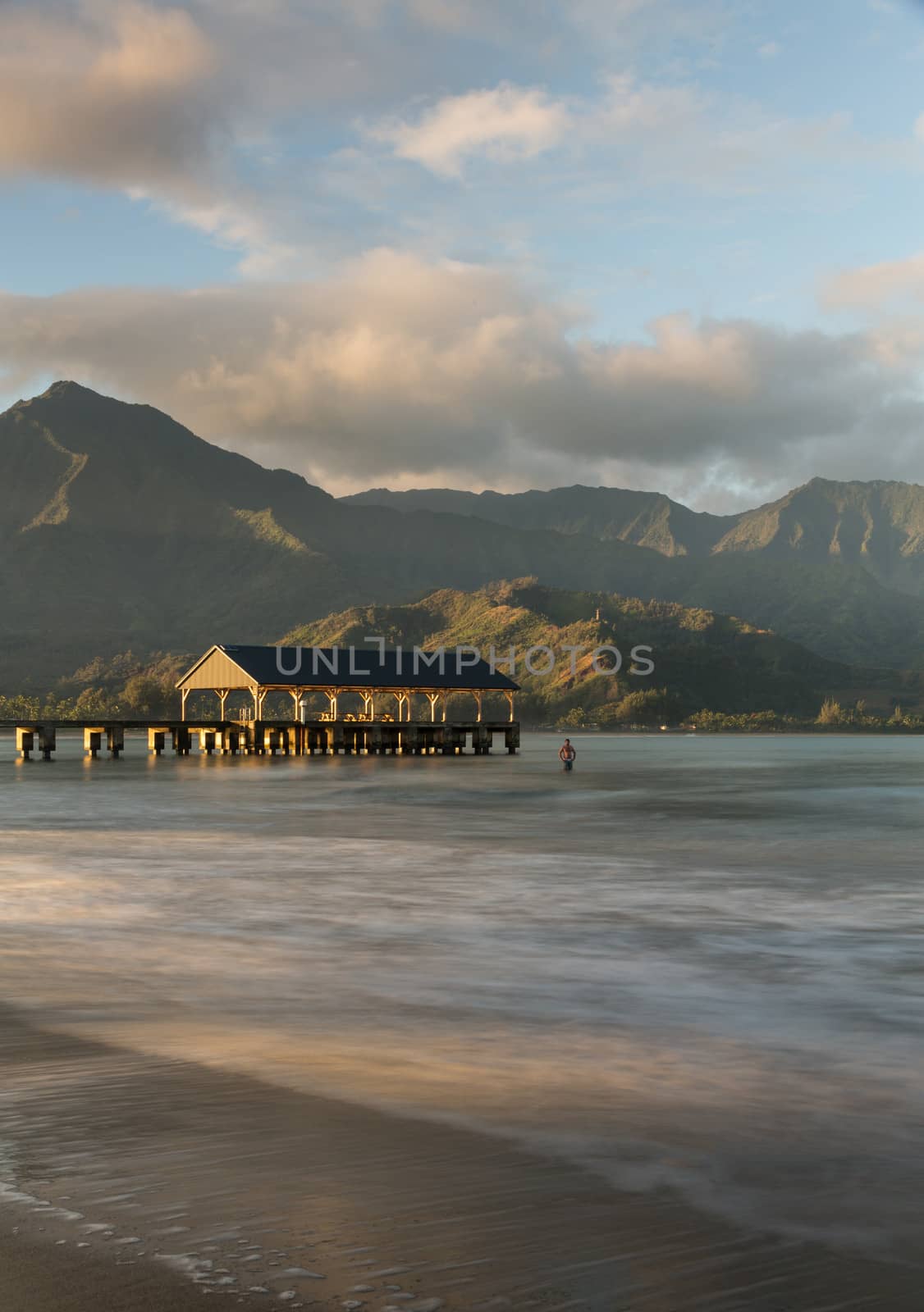 Swimmer stands in the ocean as the rising sun illuminates the peaks of Na Pali mountains over the calm bay and Hanalei Pier