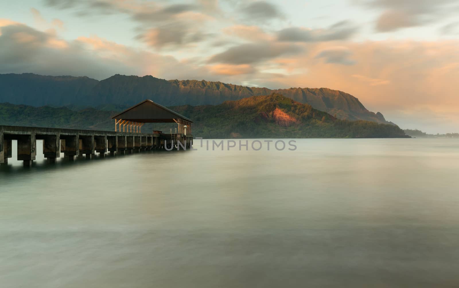 Rising sun illuminates the peaks of Na Pali mountains over the calm bay and Hanalei Pier in long exposure photo