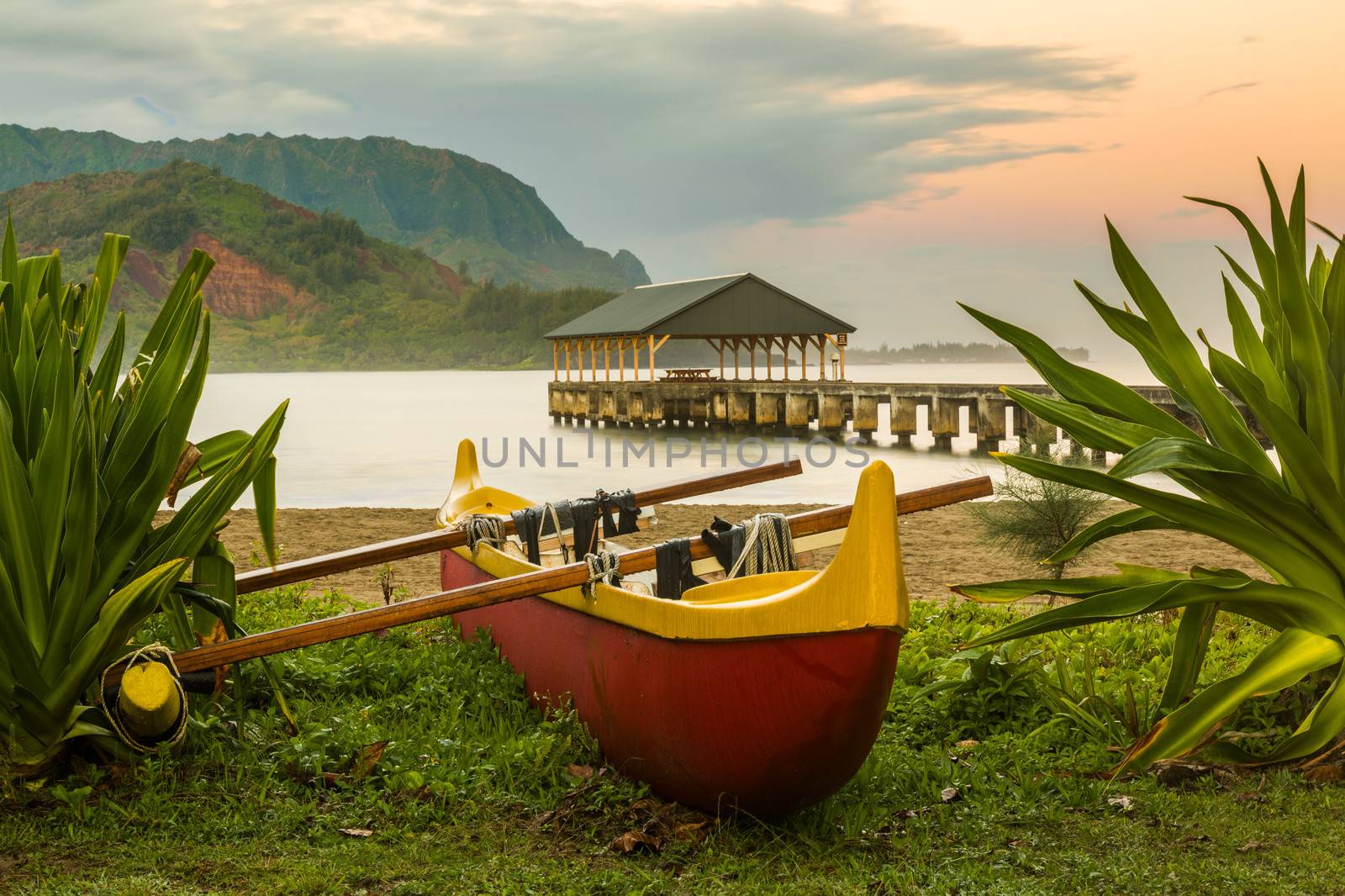 Red and yellow hawaiian canoe with outrigger on the beach at Hanalei pier at dawn as the sun lights the sky over Na Pali mountains